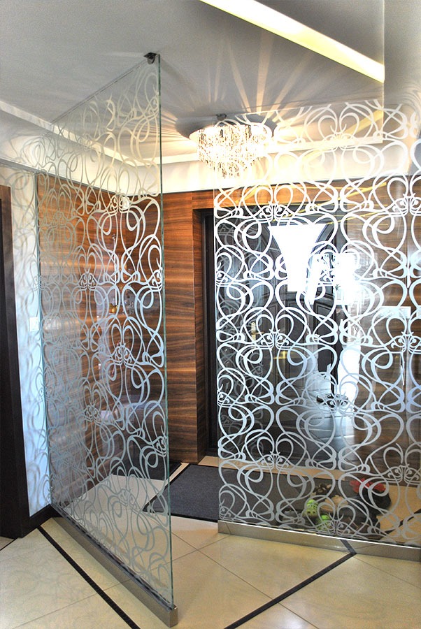 Dwie szklane ścianki działowe z ozdobnym, abstrakcyjnym wzorem oddzielają przestrzeń w jasnym wnętrzu, widoczny żyrandol kryształowy i fragment drewnianej ściany.