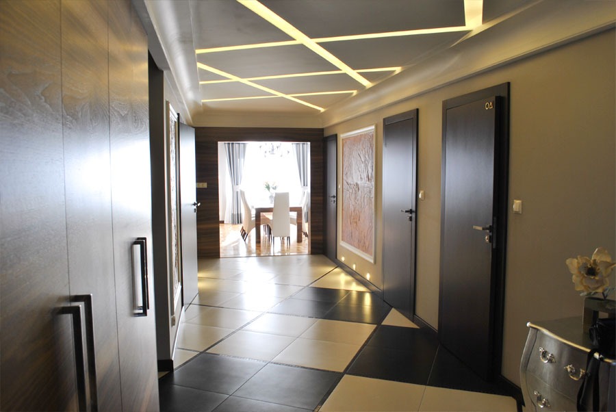 Nowoczesny korytarz z podświetlanym sufitem w geometryczne wzory, czarno-białą podłogą w szachownicę i ciemnymi drzwiami z numeracją, widok na jadalnię.