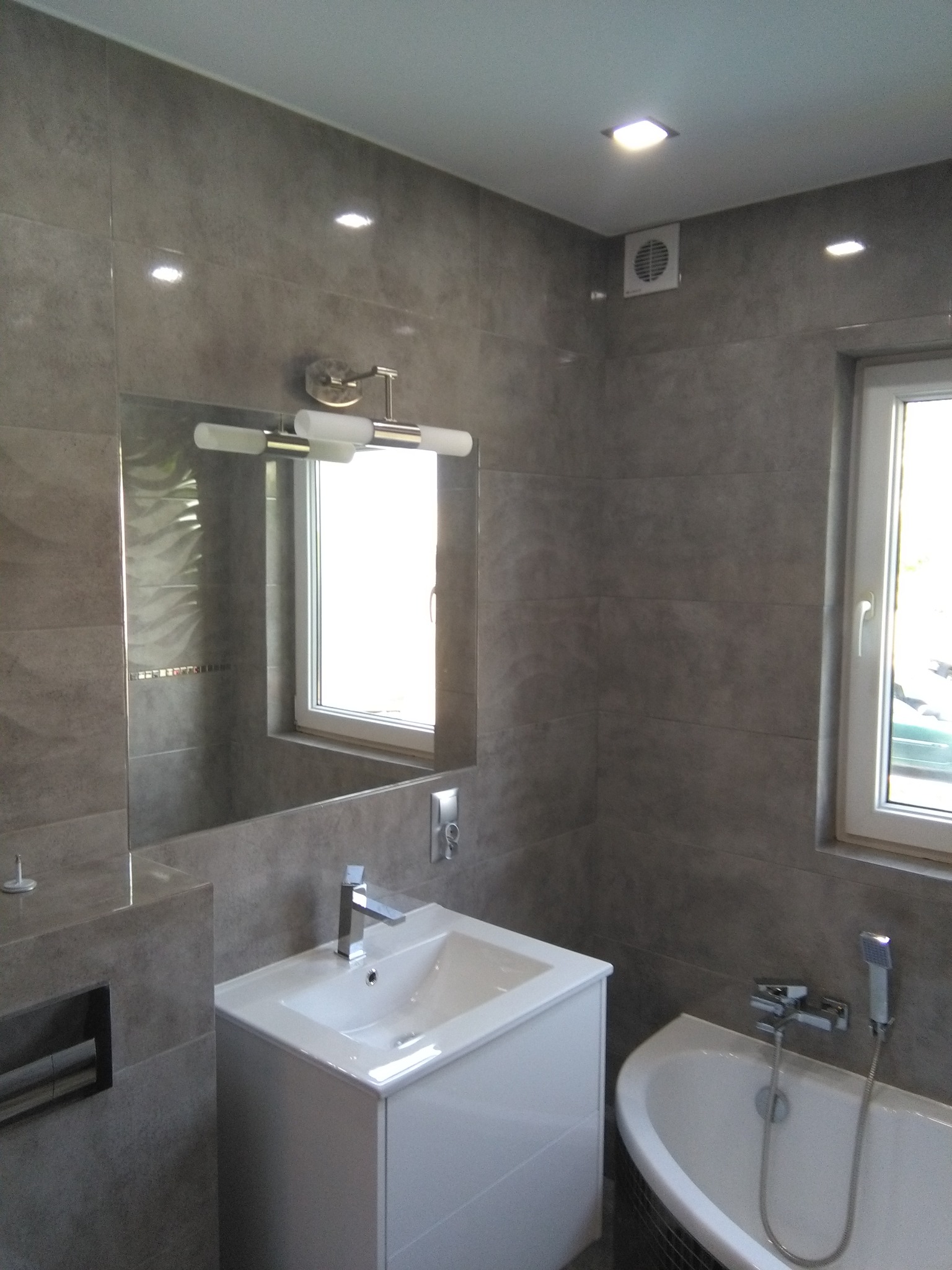 Nowoczesna łazienka z szarymi płytkami, białymi meblami, wanną narożną i dużym lustrem z oświetleniem punktowym.