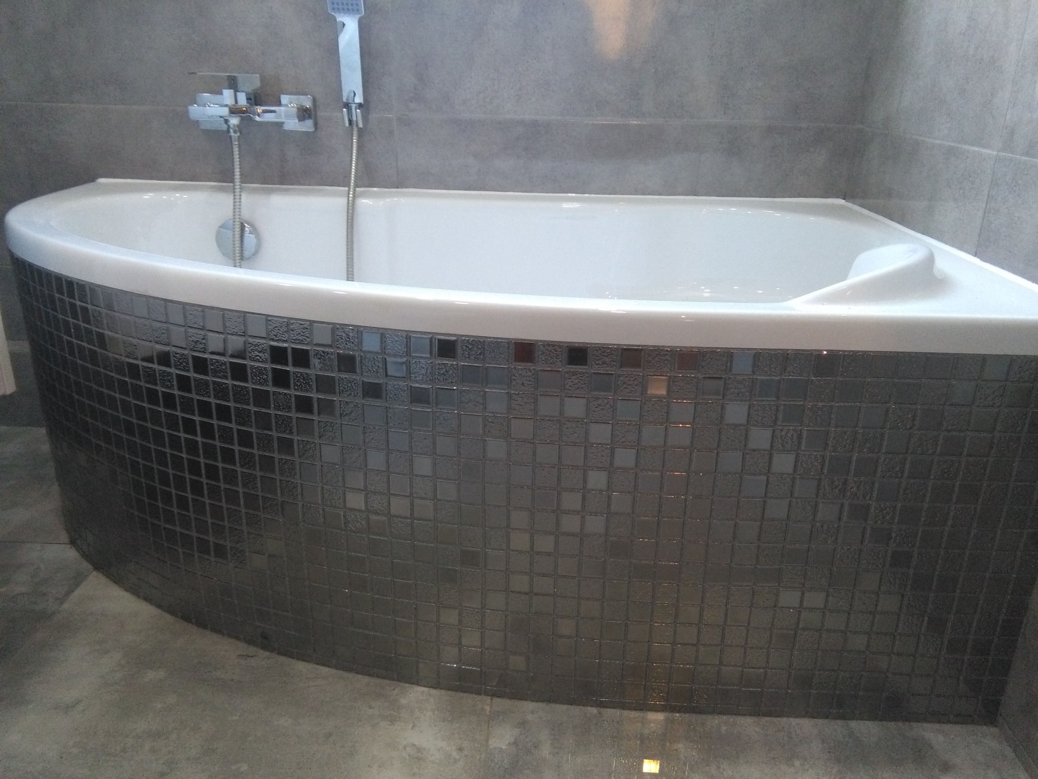 Biała, narożna wanna z obudową wykończoną mozaiką z szarych, błyszczących płytek, zamontowana w łazience z szarymi ścianami i podłogą.