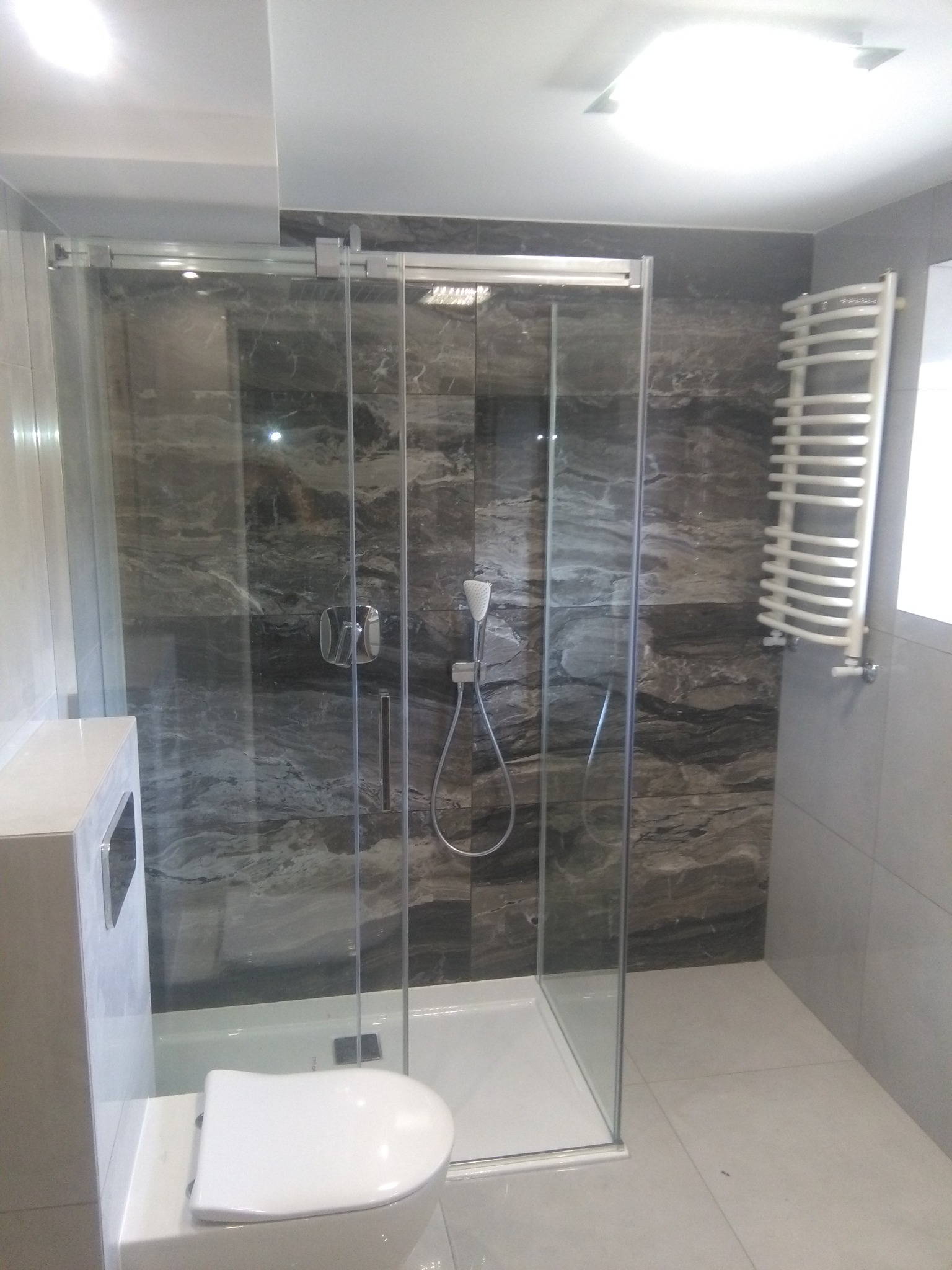 Nowoczesna łazienka z kabiną prysznicową ze szklanymi drzwiami przesuwnymi, marmurowe płytki na ścianach w odcieniach szarości, biały grzejnik drabinkowy i podwieszana toaleta.