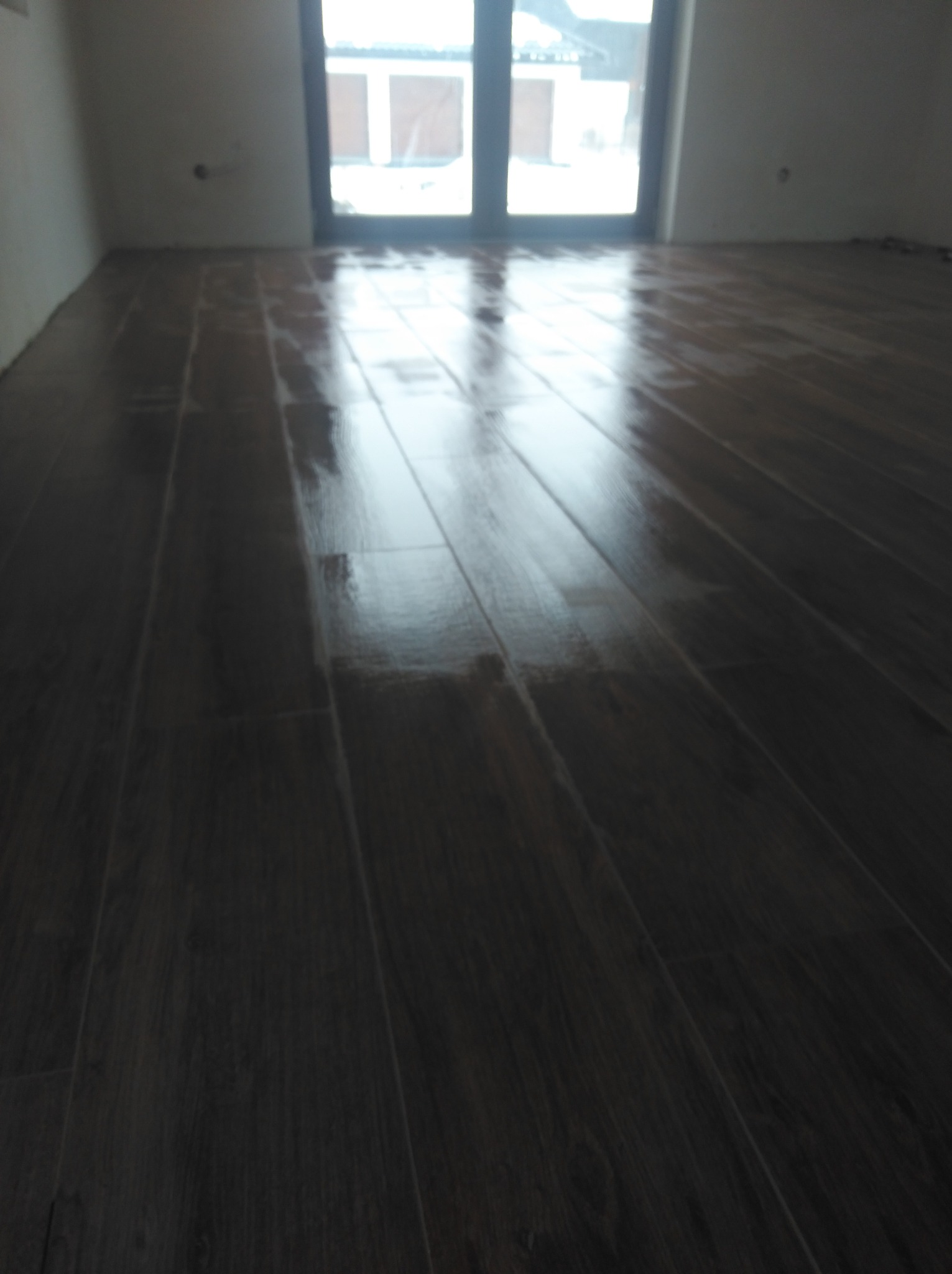 Świeżo położona, ciemnobrązowa podłoga z paneli, odbijająca światło z okna balkonowego, w surowym, białym pomieszczeniu.