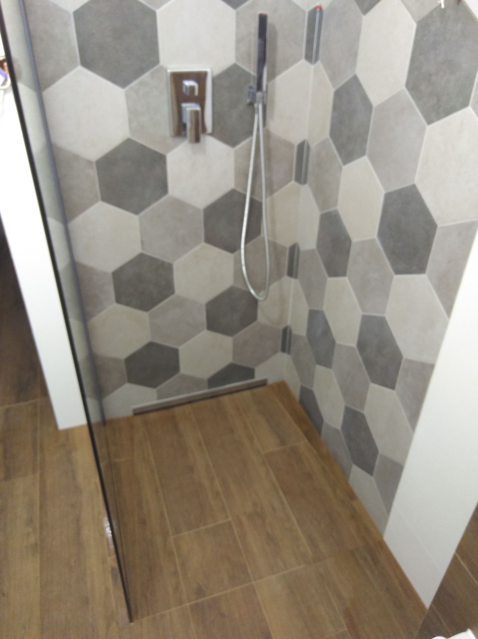Nowoczesna kabina prysznicowa z heksagonalnymi płytkami w odcieniach szarości i beżu, drewnopodobna podłoga, minimalistyczna armatura.