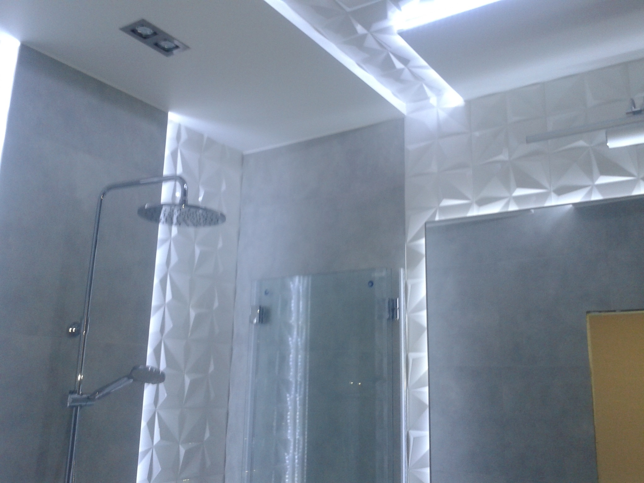 Nowoczesna łazienka z szarymi płytkami i białą trójwymiarową ścianą, szklaną kabiną prysznicową i deszczownicą, oświetlona wbudowanymi halogenami i listwą LED.