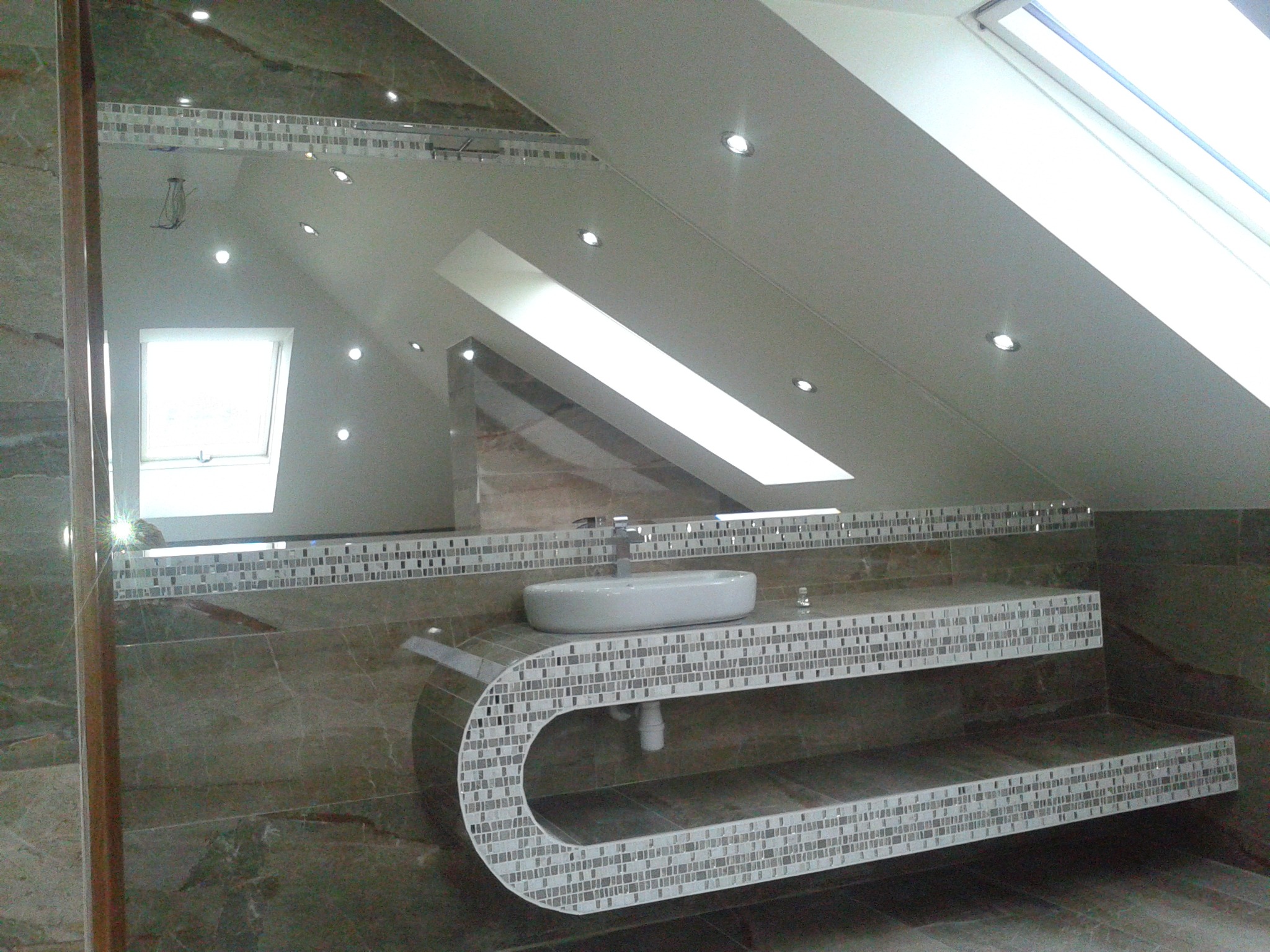 Nowoczesna łazienka na poddaszu z marmurową ścianą i blatem z mozaiki, okrągła umywalka nablatowa, oświetlenie punktowe w skosach sufitu.