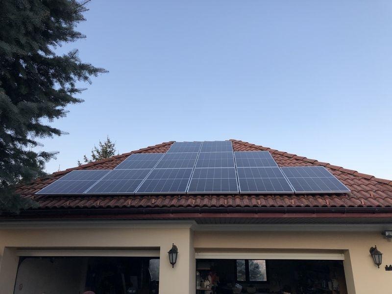 Panele słoneczne zamontowane na dachu domu z czerwoną dachówką, widok z dołu, część garażu widoczna poniżej.