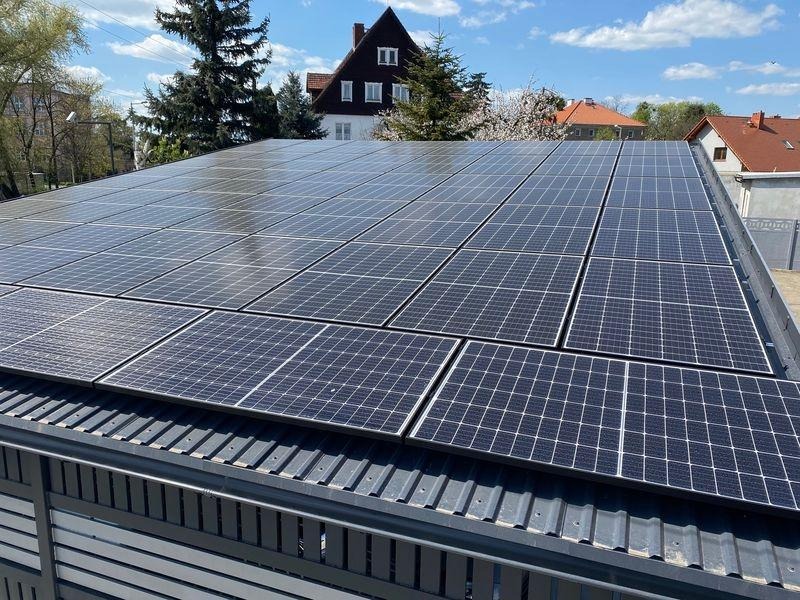 Instalacja paneli słonecznych na dachu garażu z blachy trapezowej, widok z góry, w tle budynki mieszkalne i drzewa pod błękitnym niebem.