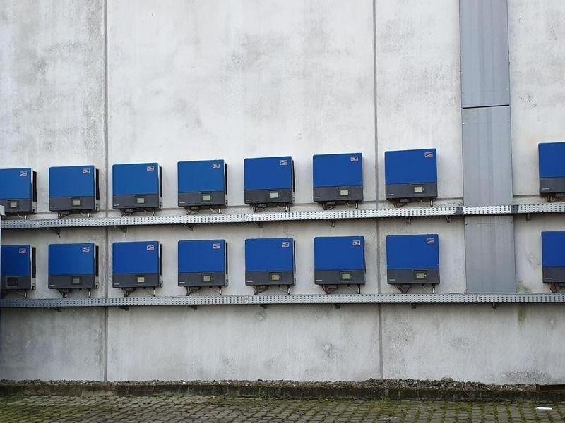 Rząd niebieskich inwerterów fotowoltaicznych zamontowanych na szarej, betonowej ścianie budynku przemysłowego, widoczne kable i metalowe prowadnice.