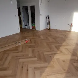 Jasny parkiet ułożony we wzór jodełki w pokoju w trakcie remontu; paczki z panelami leżą na podłodze, ściany przygotowane do malowania.