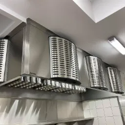 Profesjonalny okap kuchenny marki Jeven ze stali nierdzewnej, zamontowany w kuchni z białymi kafelkami, oświetlony lampą sufitową.