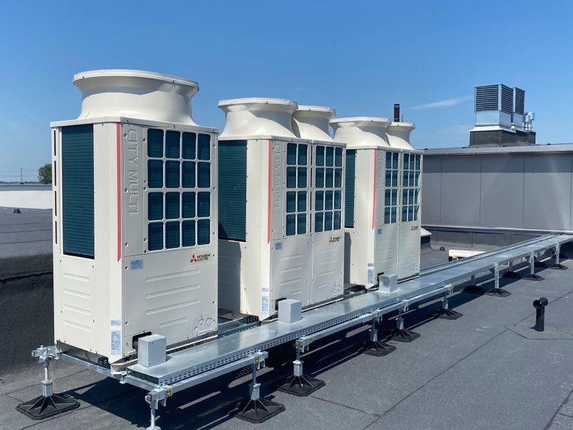 Cztery jednostki zewnętrzne systemu klimatyzacji CITY MULTI firmy Mitsubishi Electric, zamontowane na dachu budynku na specjalnej konstrukcji wsporczej, widoczne w słoneczny dzień.