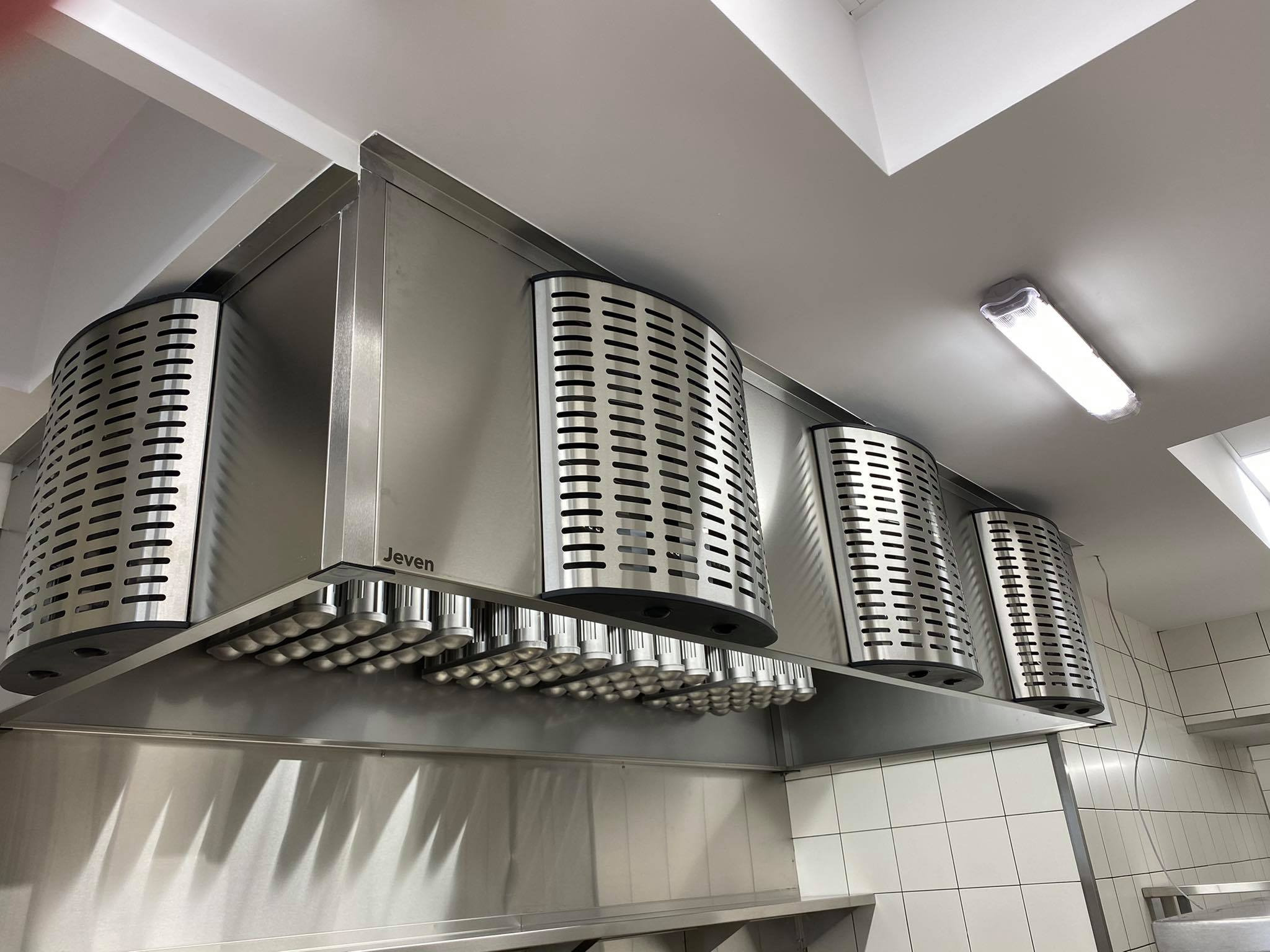 Profesjonalny okap kuchenny marki Jeven ze stali nierdzewnej, zamontowany w kuchni z białymi kafelkami, oświetlony lampą sufitową.