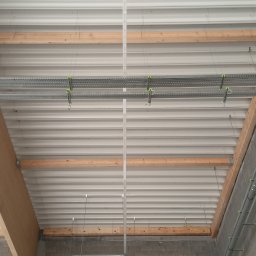 Andrzej Pawelczak - Montaż instalacji pod stropem z blachy trapezowej. Widoczne korytka kablowe, rury i elementy konstrukcyjne. Surowy stan wykończenia wnętrza.