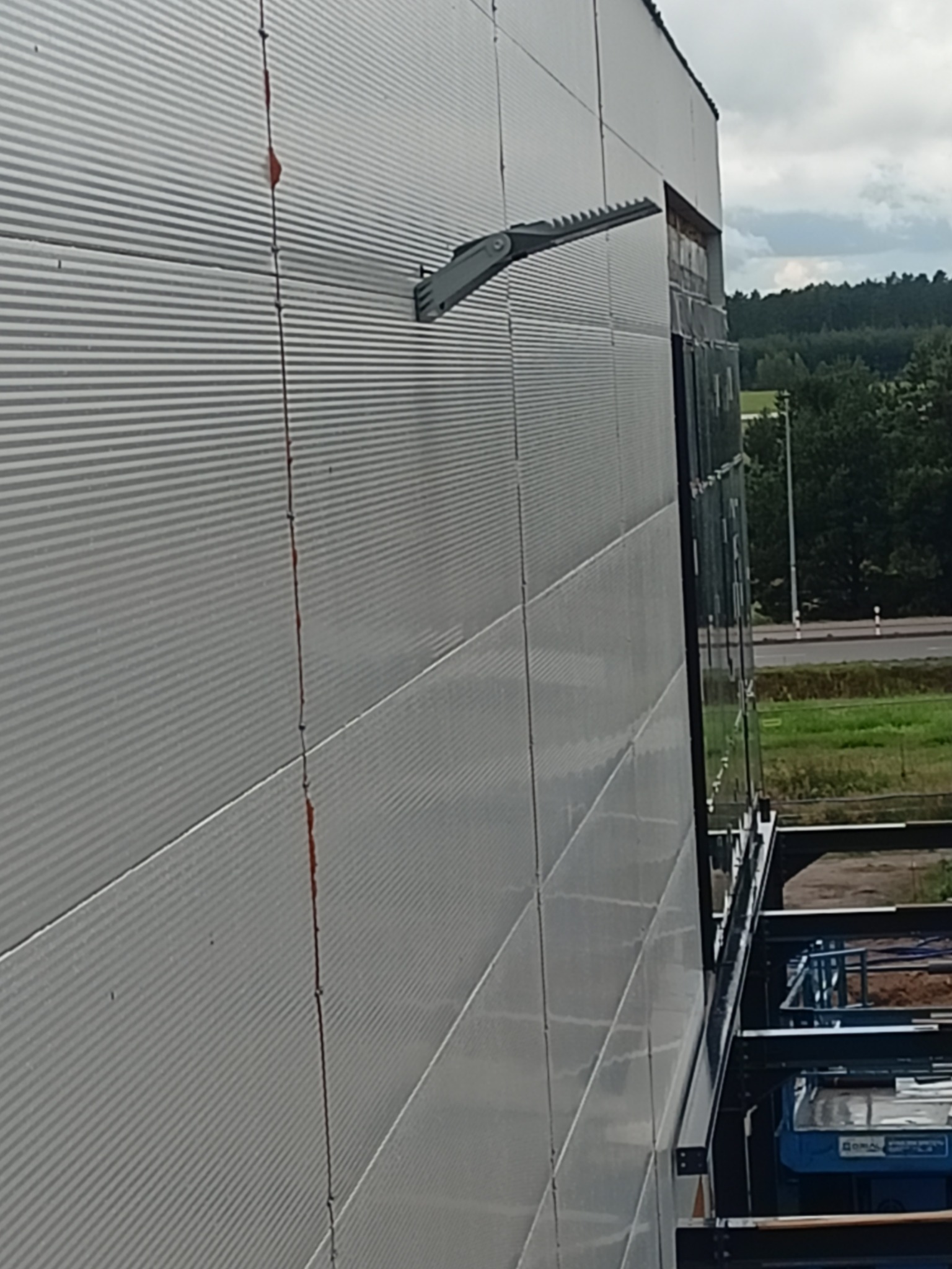 Szara, modułowa elewacja hali przemysłowej z zamontowaną lampą LED. Widoczne fragmenty konstrukcji i otoczenia.