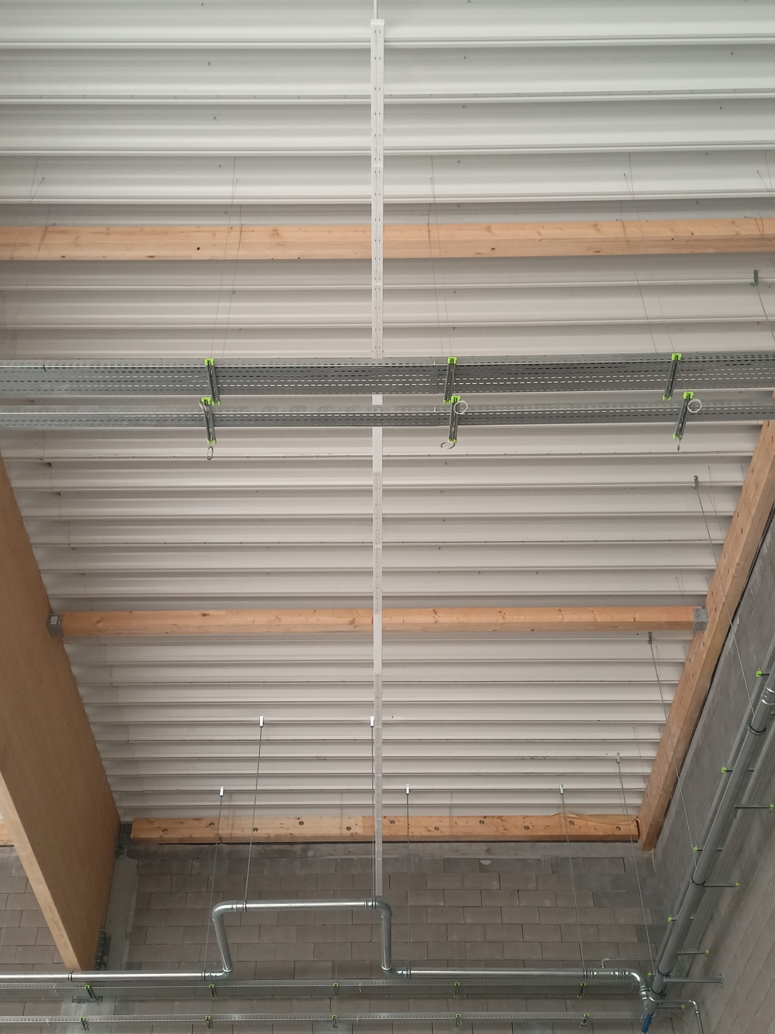 Montaż instalacji pod stropem z blachy trapezowej. Widoczne korytka kablowe, rury i elementy konstrukcyjne. Surowy stan wykończenia wnętrza.