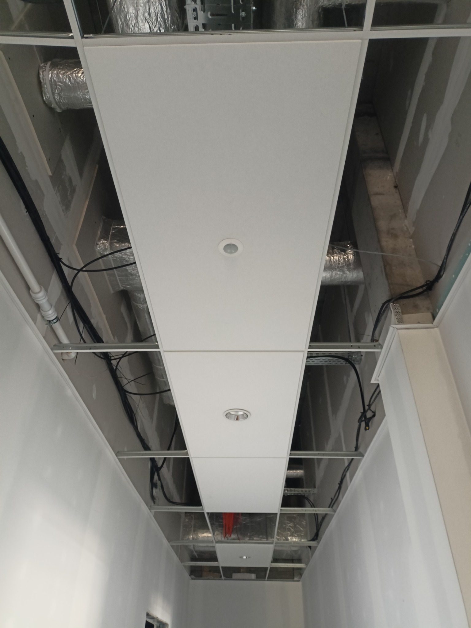 Biały sufit podwieszany z oświetleniem punktowym i widocznymi instalacjami wentylacyjnymi oraz elektrycznymi w przestrzeni między sufitem a stropem.