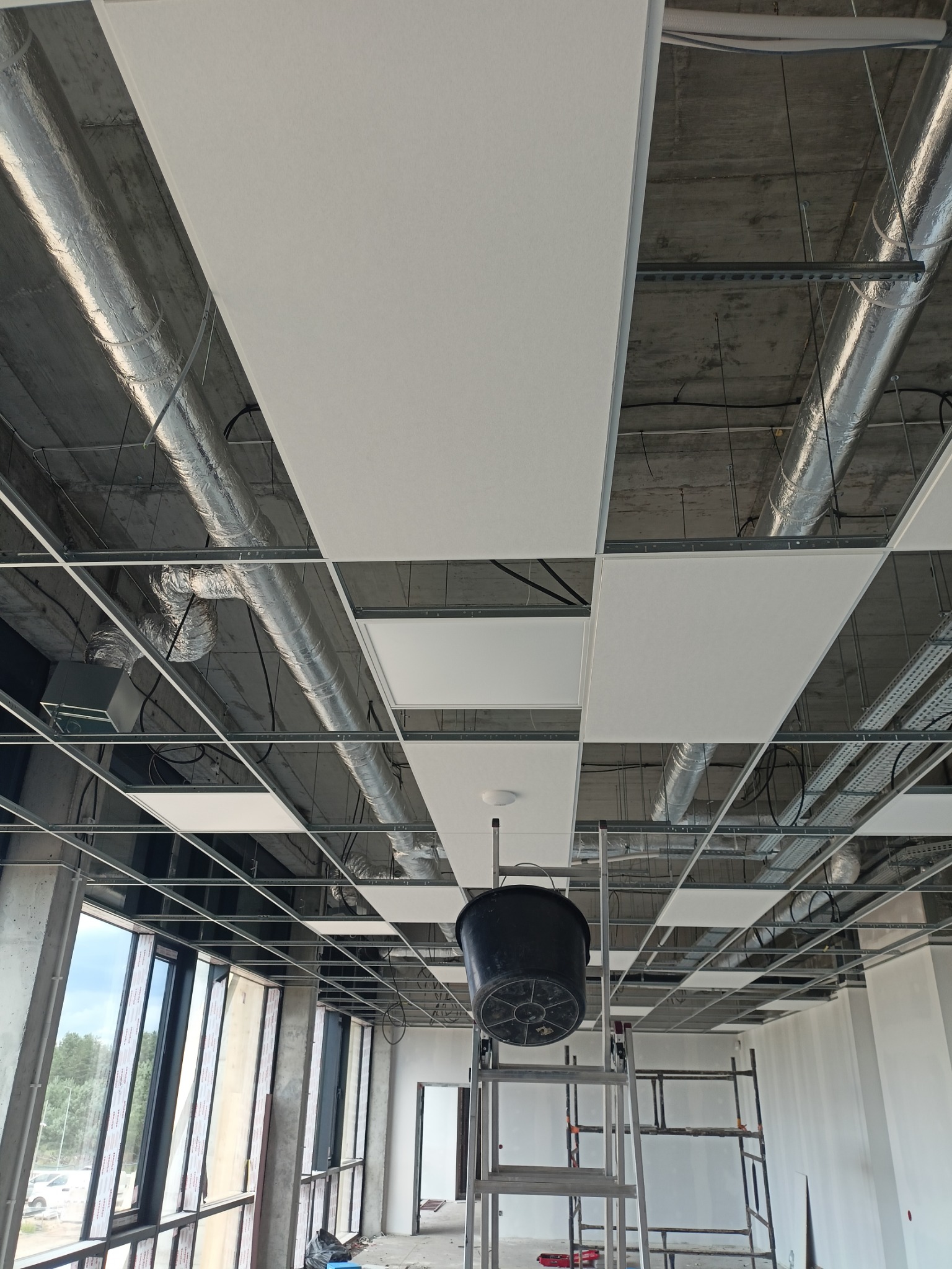 Montaż sufitu podwieszanego kasetonowego z widocznymi instalacjami wentylacyjnymi. Na pierwszym planie drabina z wiadrem, w tle rusztowanie. Jasne wnętrze biurowe.