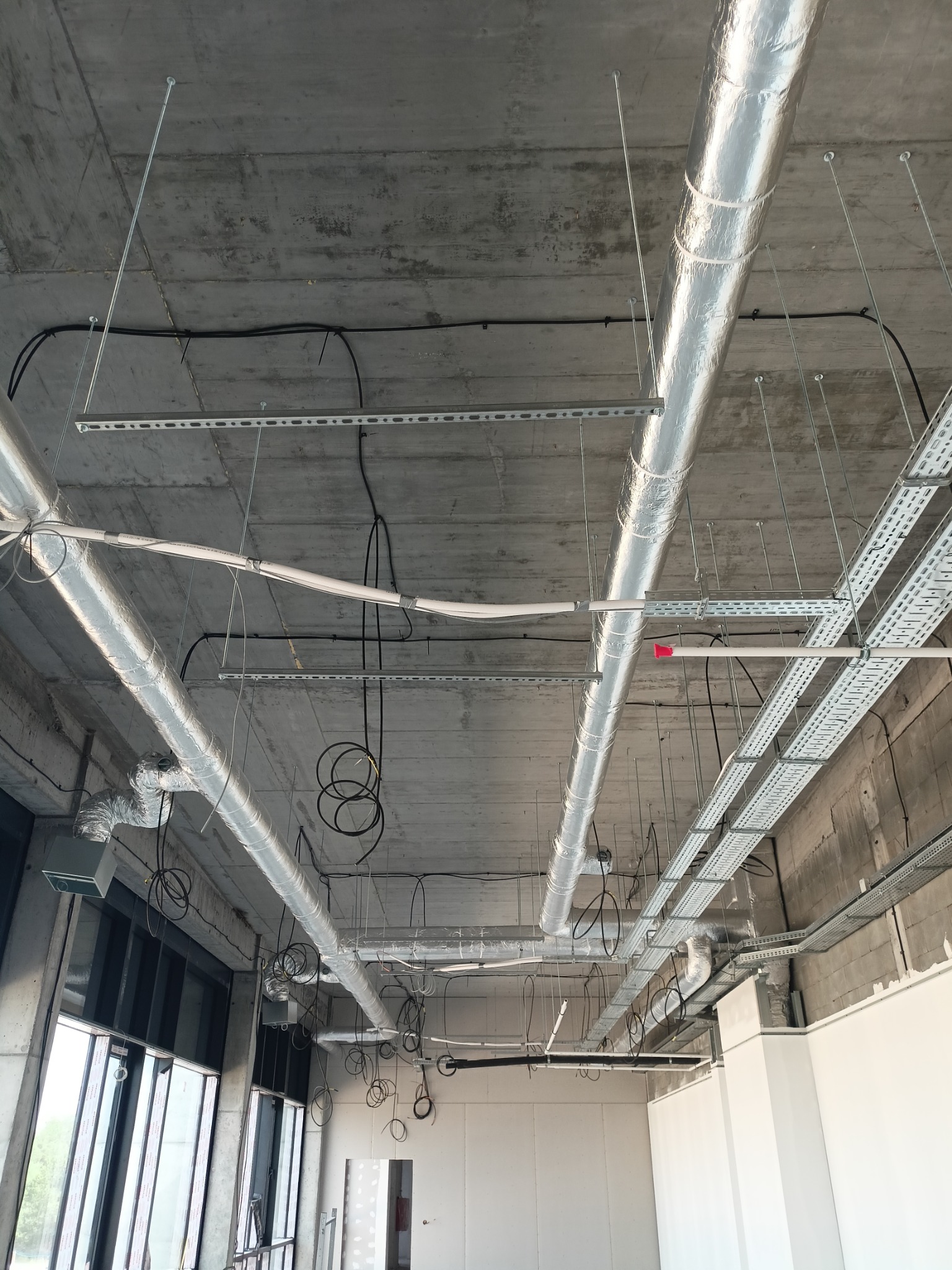 Widok sufitu z instalacjami: rury wentylacyjne w srebrnej izolacji, koryta kablowe z przewodami, betonowy strop. Widoczne okna i fragment ścian. Instalacje w stanie surowym.