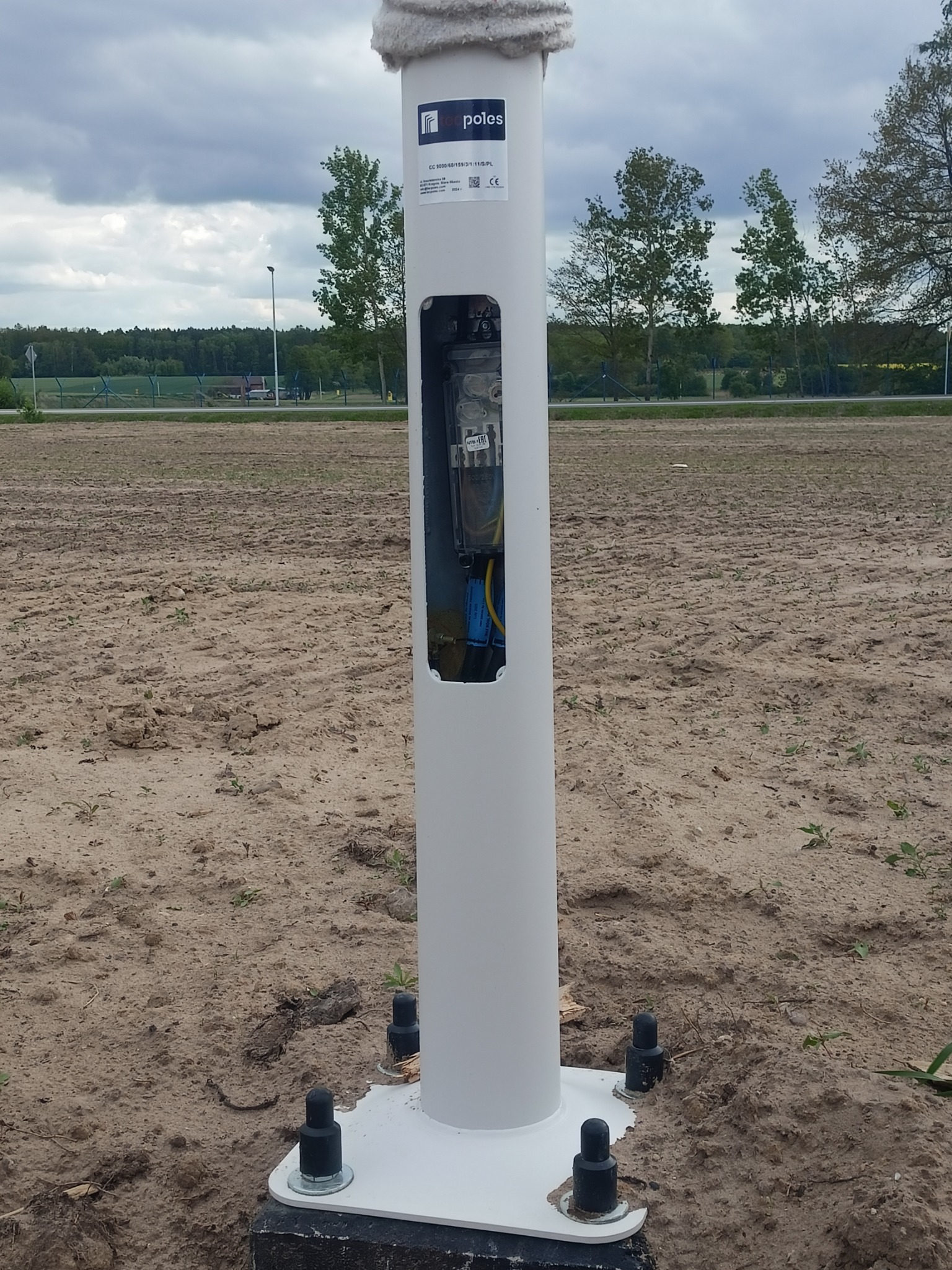 Biała słupkowa obudowa z otwartym panelem elektrycznym na zewnątrz, na tle pola i drzew. Widoczne okablowanie i elementy instalacji.