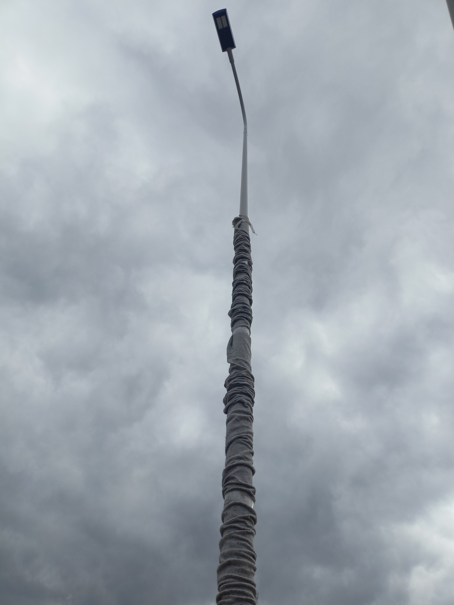 Słup oświetleniowy z lampą LED, owinięty materiałem izolacyjnym, na tle pochmurnego nieba. Nietypowe zabezpieczenie słupa przed warunkami atmosferycznymi.