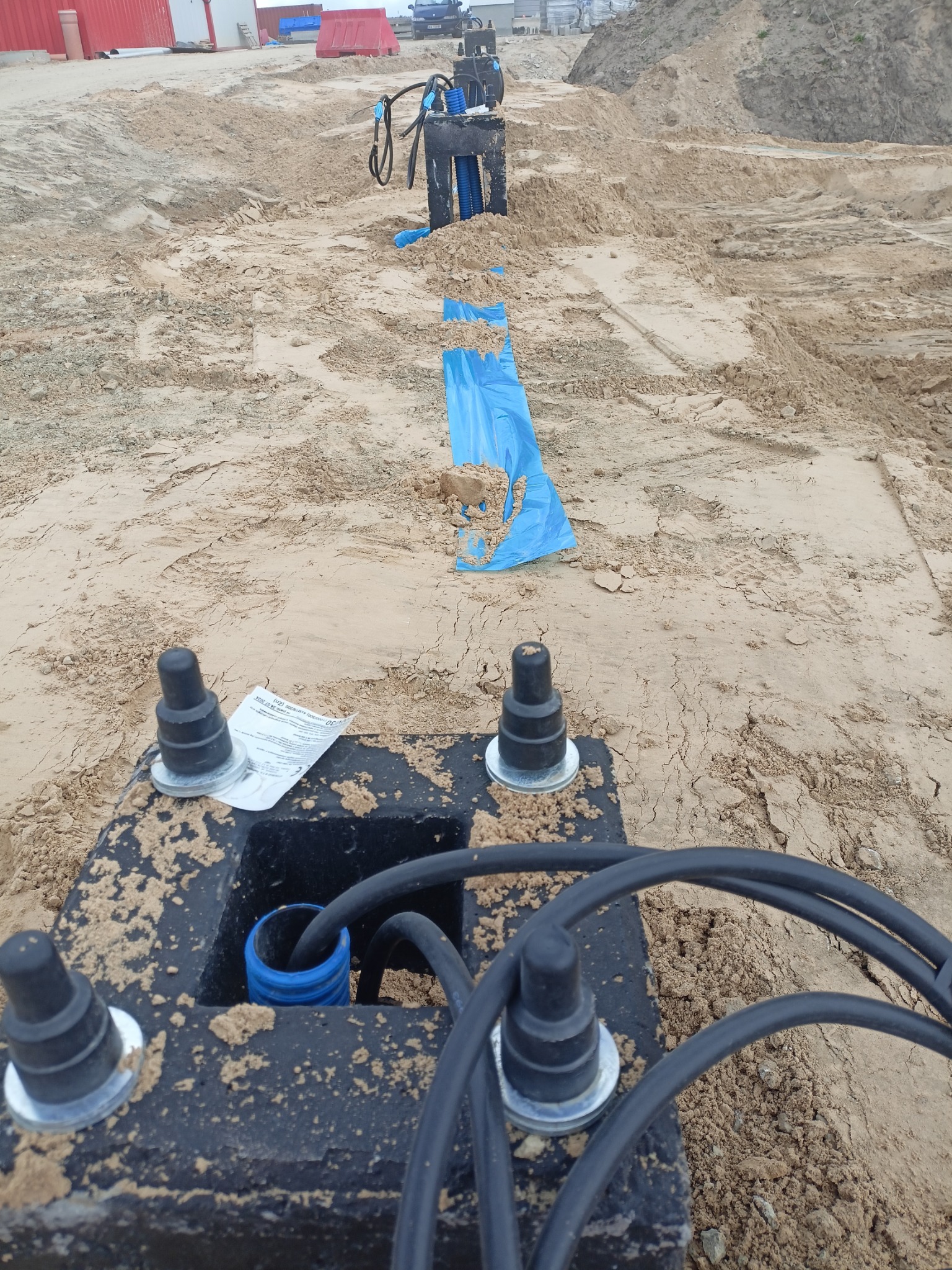 Wkopane w piasek czarne studzienki kablowe z niebieskimi rurami karbowanymi i kablami. Widoczne elementy instalacji odgromowej z papierową instrukcją.