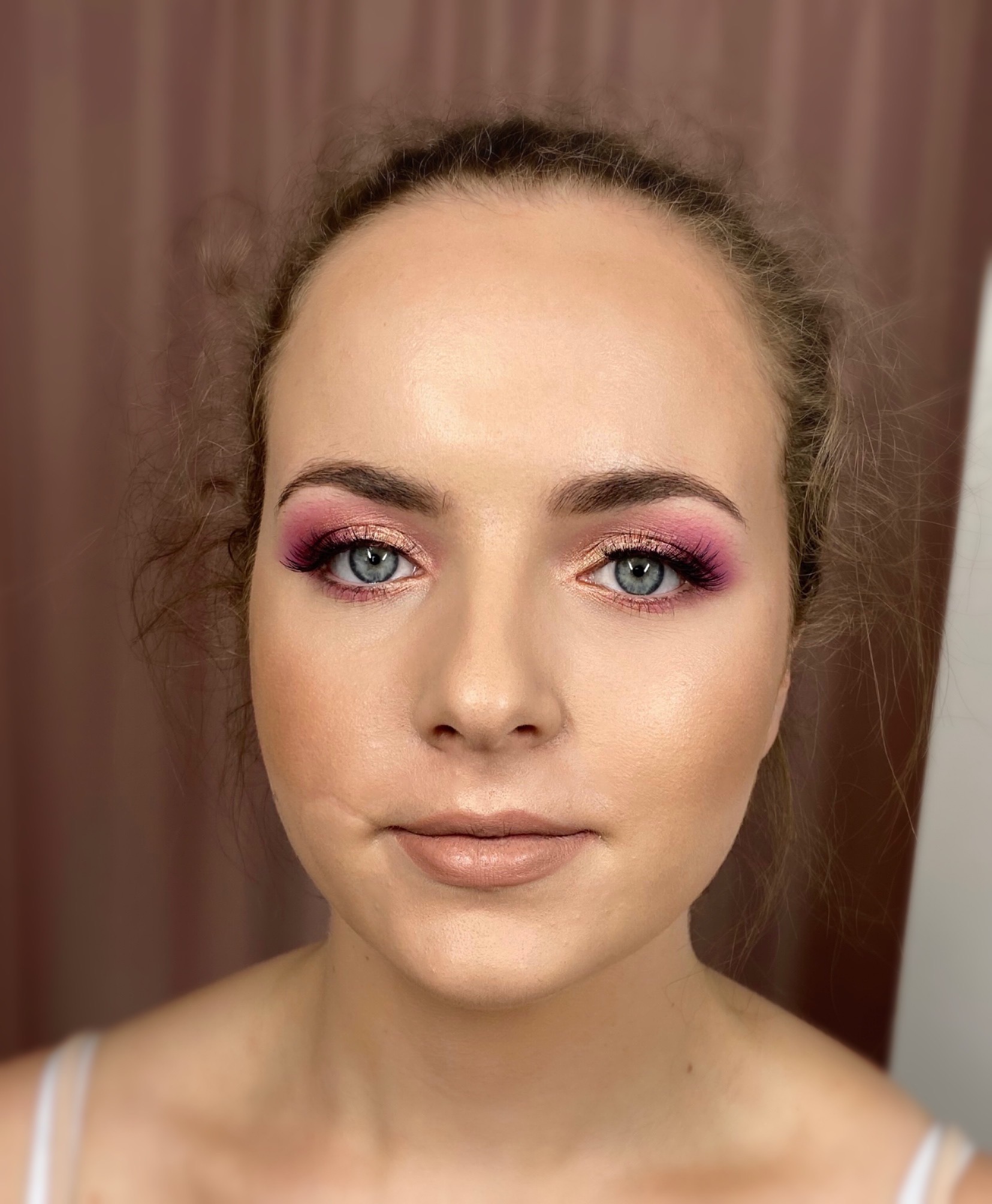 Portret kobiety z delikatnym, różowo-złotym cieniowaniem powiek i sztucznymi rzęsami, podkreślającym błękitne oczy. Usta w odcieniu nude z błyszczącym wykończeniem.
