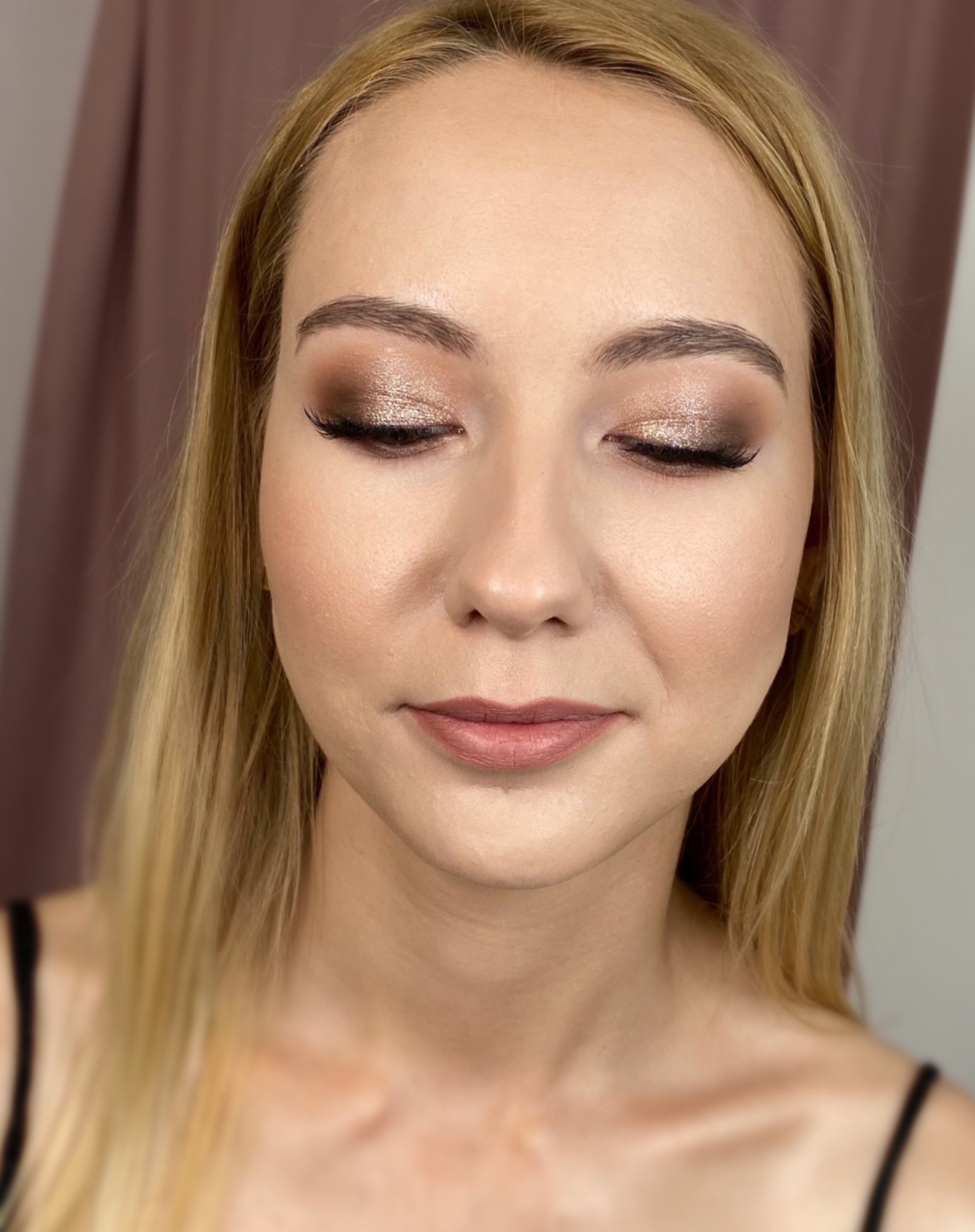 Portret kobiety z zamkniętymi oczami, prezentujący makijaż w odcieniach brązu i złota z wyraźnie podkreślonymi rzęsami i ustami w kolorze nude.