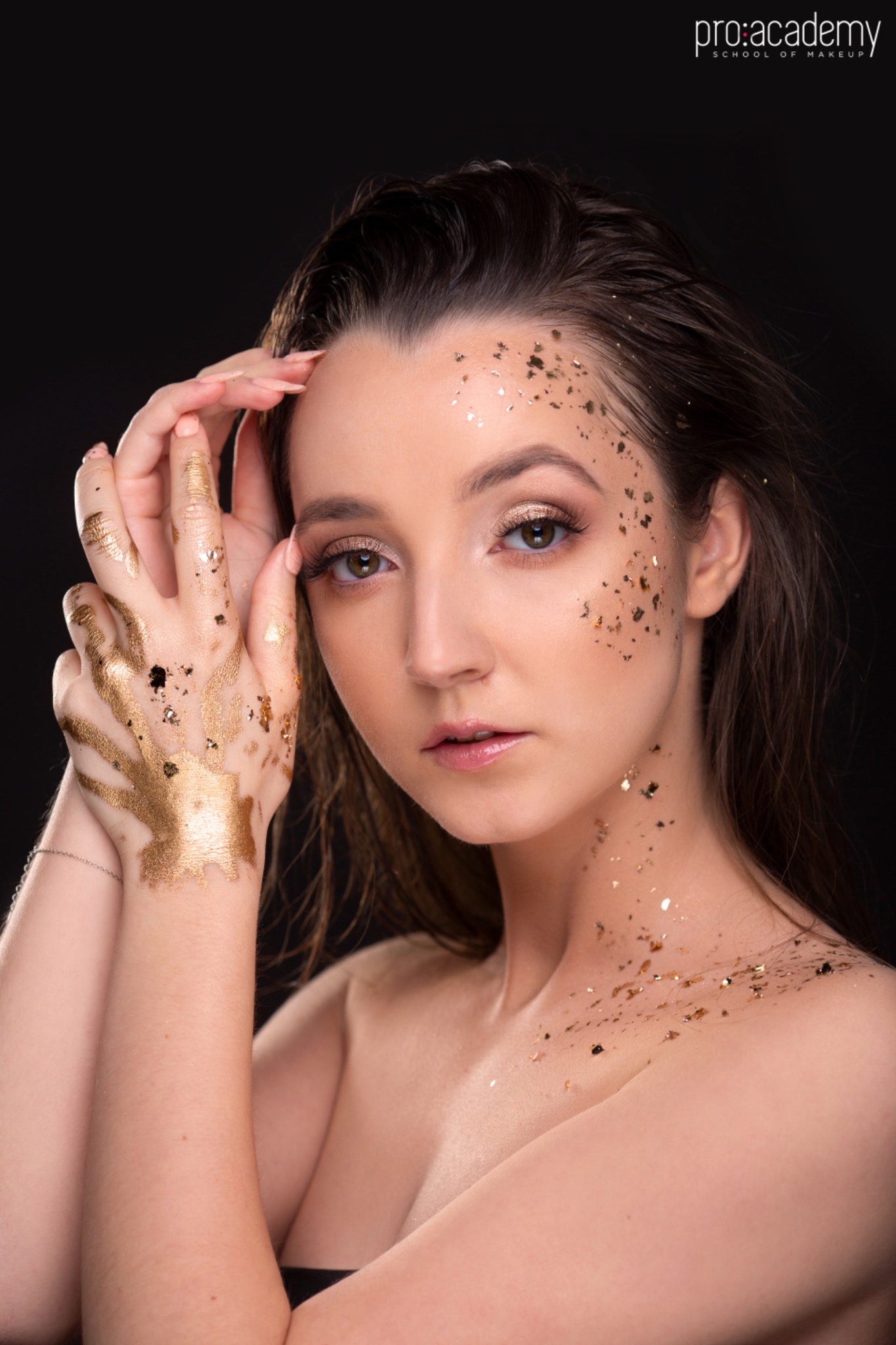 Portret kobiety z artystycznym zdobieniem złotymi płatkami na twarzy i dłoniach, z delikatnym makijażem oczu i ust, na ciemnym tle.