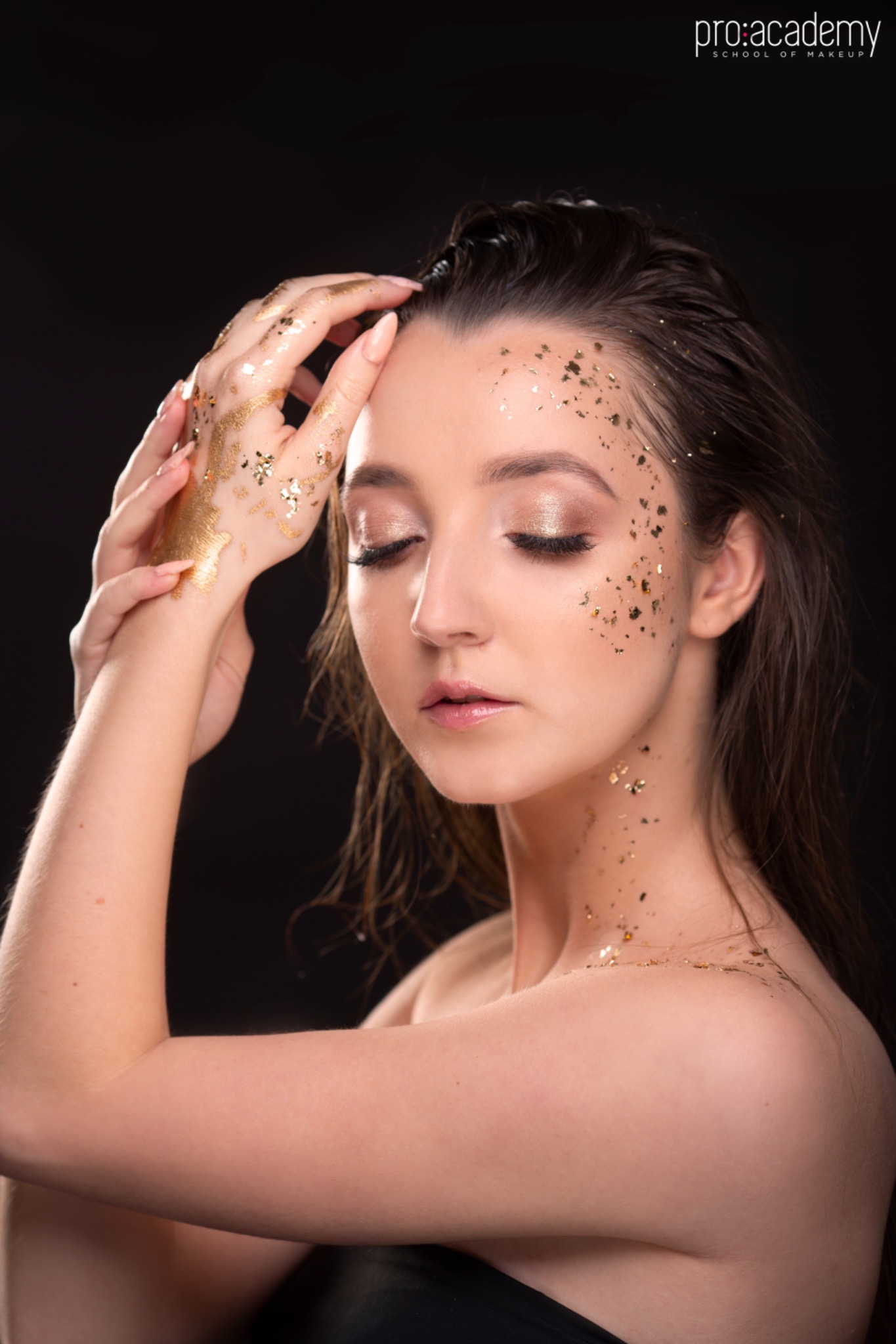 Portret kobiety z zamkniętymi oczami, z artystycznym zdobieniem twarzy i dłoni złotymi płatkami, na ciemnym tle.