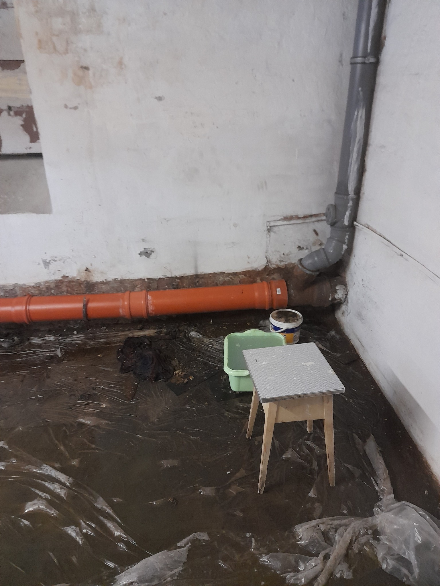 Pomieszczenie w trakcie prac remontowych instalacji hydraulicznej: widoczne rury PCV, wiadro, stołek i folia ochronna na podłodze.
