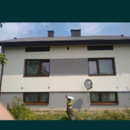 Dwukondygnacyjny dom z elewacją w odcieniach bieli i szarości, brązowe okna z firankami, antena satelitarna na ścianie, w ogr&oacute;dku dekoracja z pnia drzewa.