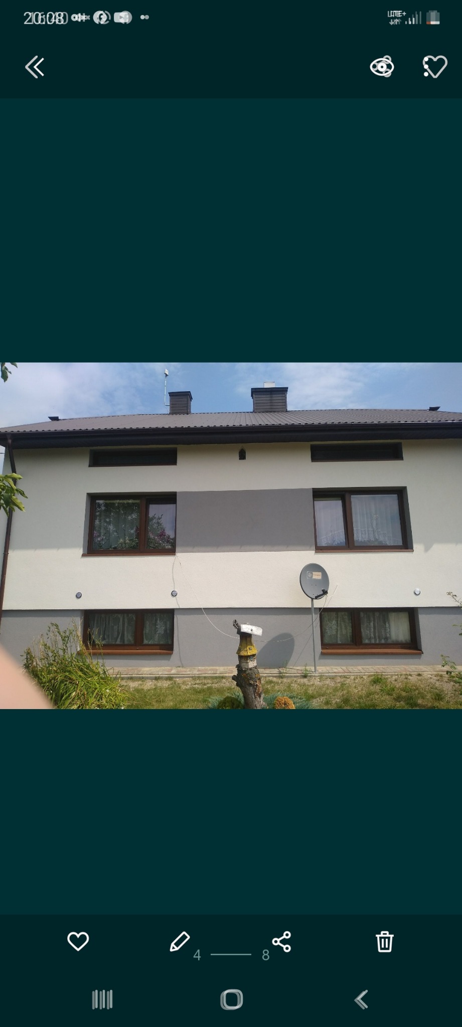 Dwukondygnacyjny dom z elewacją w odcieniach bieli i szarości, brązowe okna z firankami, antena satelitarna na ścianie, w ogródku dekoracja z pnia drzewa.