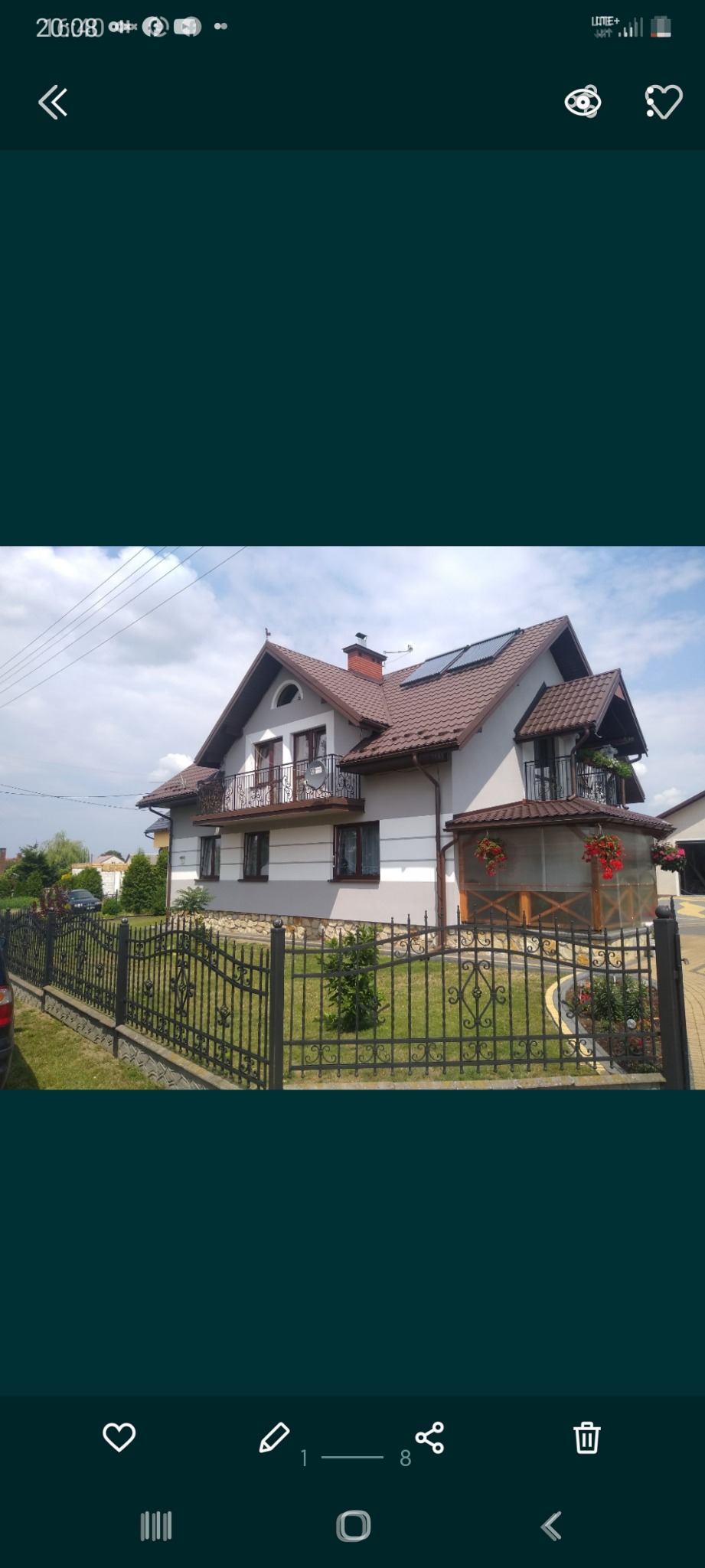 Dwupiętrowy dom z szarą elewacją, brązowym dachem, balkonami z kutymi balustradami i panelami słonecznymi na dachu, widoczny zza czarnego kutego ogrodzenia.