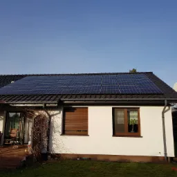 Dom jednorodzinny z panelami słonecznymi na dachu z ciemnej dachówki, widok z poziomu trawnika, antena satelitarna na ścianie bocznej, częściowo zasłonięte okno z roletą.