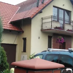 Pracownik w kombinezonie czyści czerwoną dachówkę domu jednorodzinnego za pomocą myjki ciśnieniowej, widoczna piana i woda spływająca po dachu.