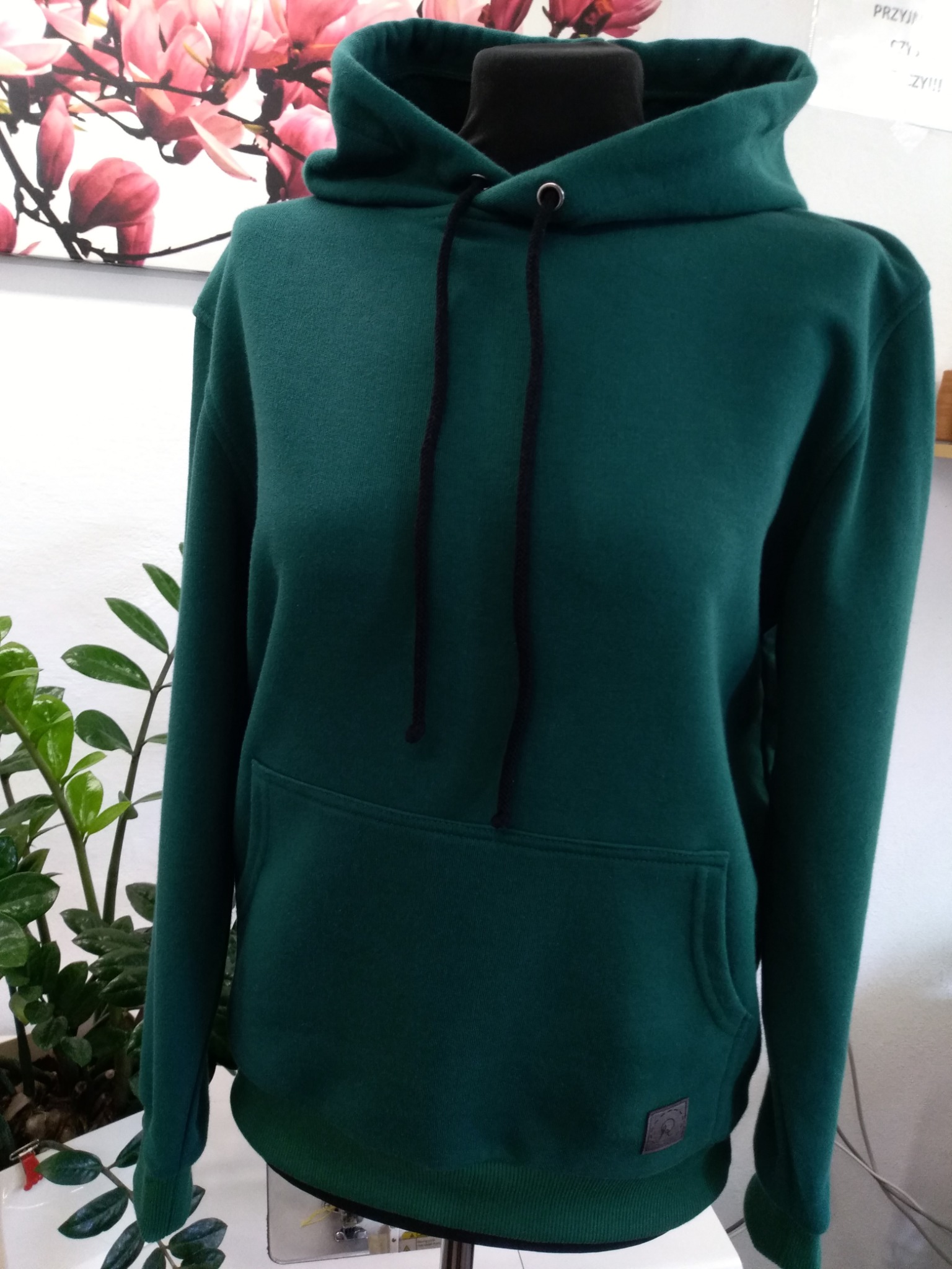 Szmaragdowa bluza z kapturem, z czarnymi sznurkami, prezentowana na manekinie na tle białej ściany z dekoracją kwiatową i zieloną rośliną doniczkową.