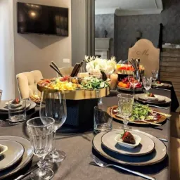 Elegancko nakryty stół z przekąskami i daniami na talerzach, kwiaty w centralnej części, telewizor na ścianie w tle