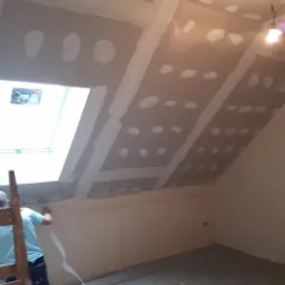 Montaż płyt gipsowo-kartonowych na poddaszu ze skosami, widoczne łączenia zaszpachlowane, pracownik na drabinie przy oknie dachowym, w tle surowy beton na podłodze.