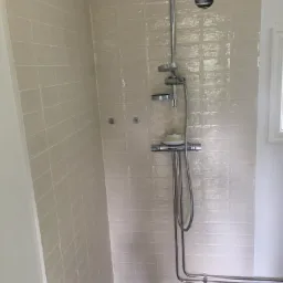 Beżowa łazienka z kwadratowymi płytkami, chromowaną armaturą prysznicową i szarą podłogą. Widoczny otwór wentylacyjny w górnej części ściany.