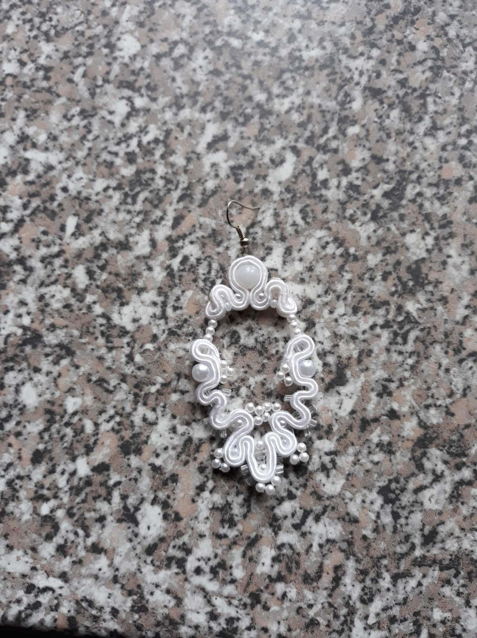 Delikatny, ręcznie robiony kolczyk sutasz w kolorze białym z perłami, leżący na marmurowym blacie.