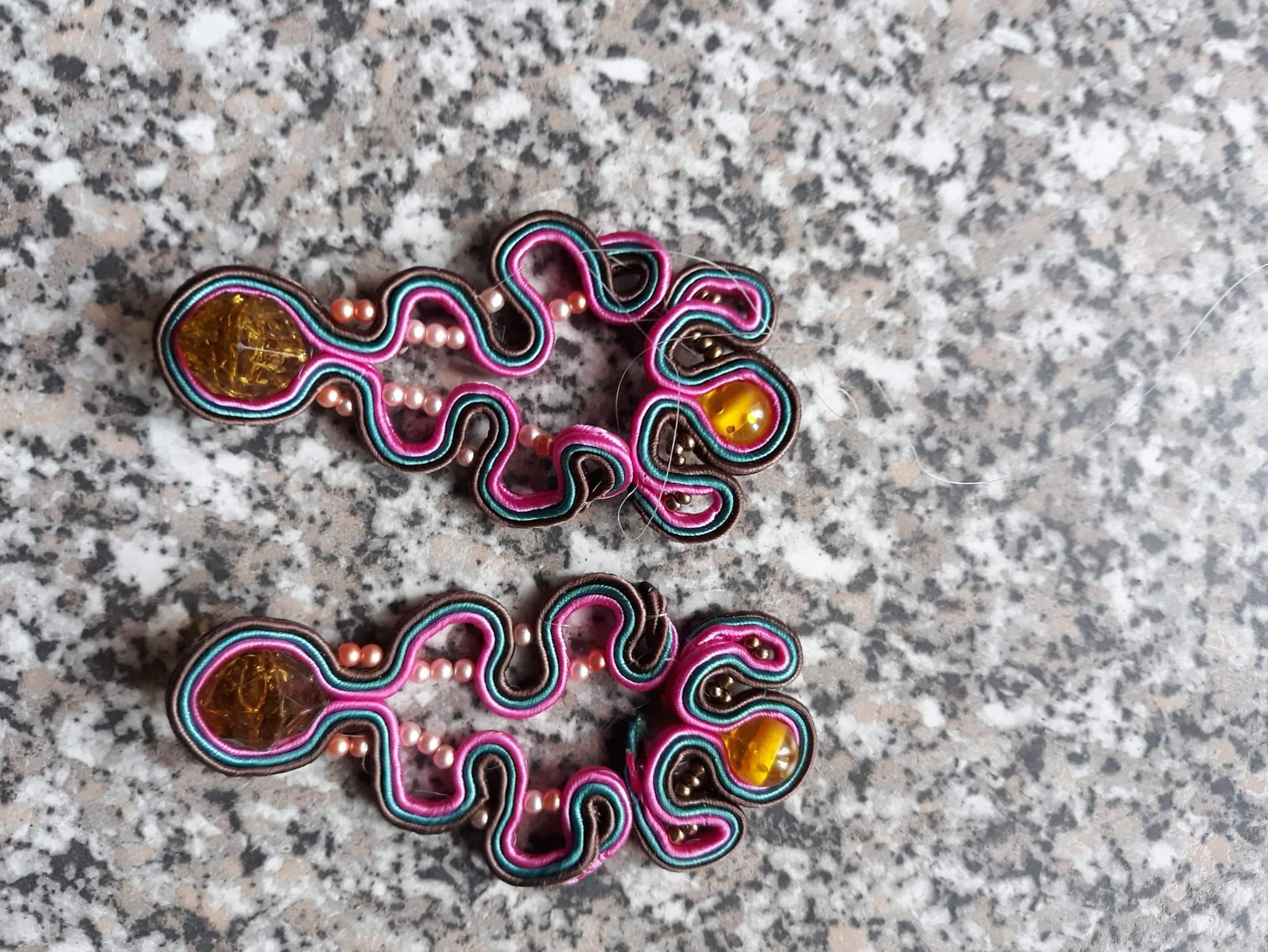 Dwa ręcznie robione kolczyki sutasz z brązowymi kamieniami, różowymi perłami i wielokolorowymi sznurkami na marmurowym tle.