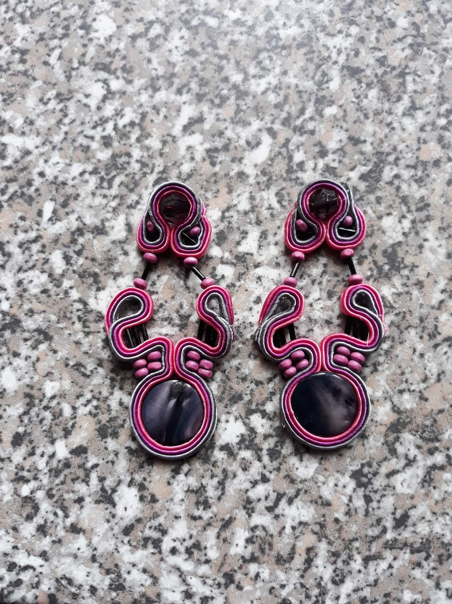 Dwa ręcznie robione kolczyki sutasz w odcieniach różu, szarości i czerni, z okrągłymi kamieniami, leżące na marmurowej powierzchni.