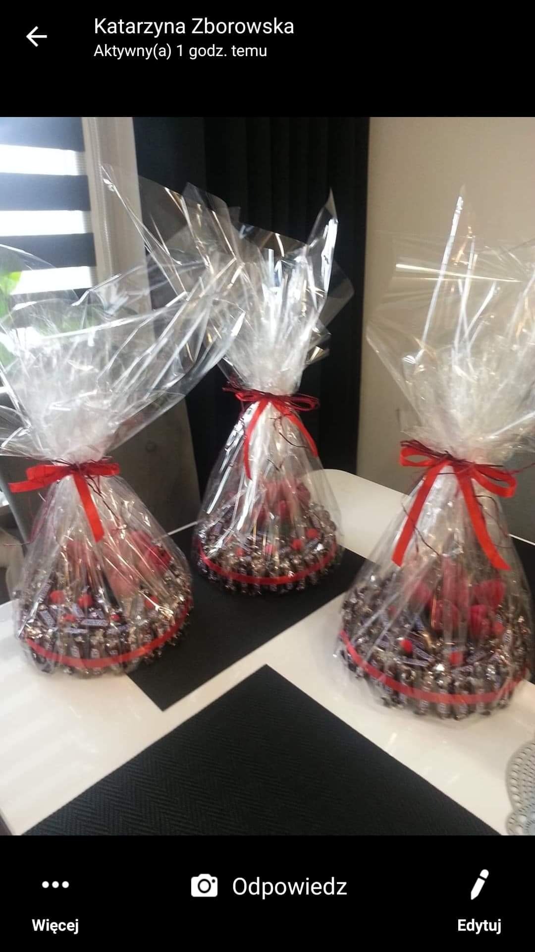 Trzy okrągłe kompozycje słodyczy, owinięte w przezroczysty celofan i przewiązane czerwoną wstążką, ustawione na czarno-białym stole.