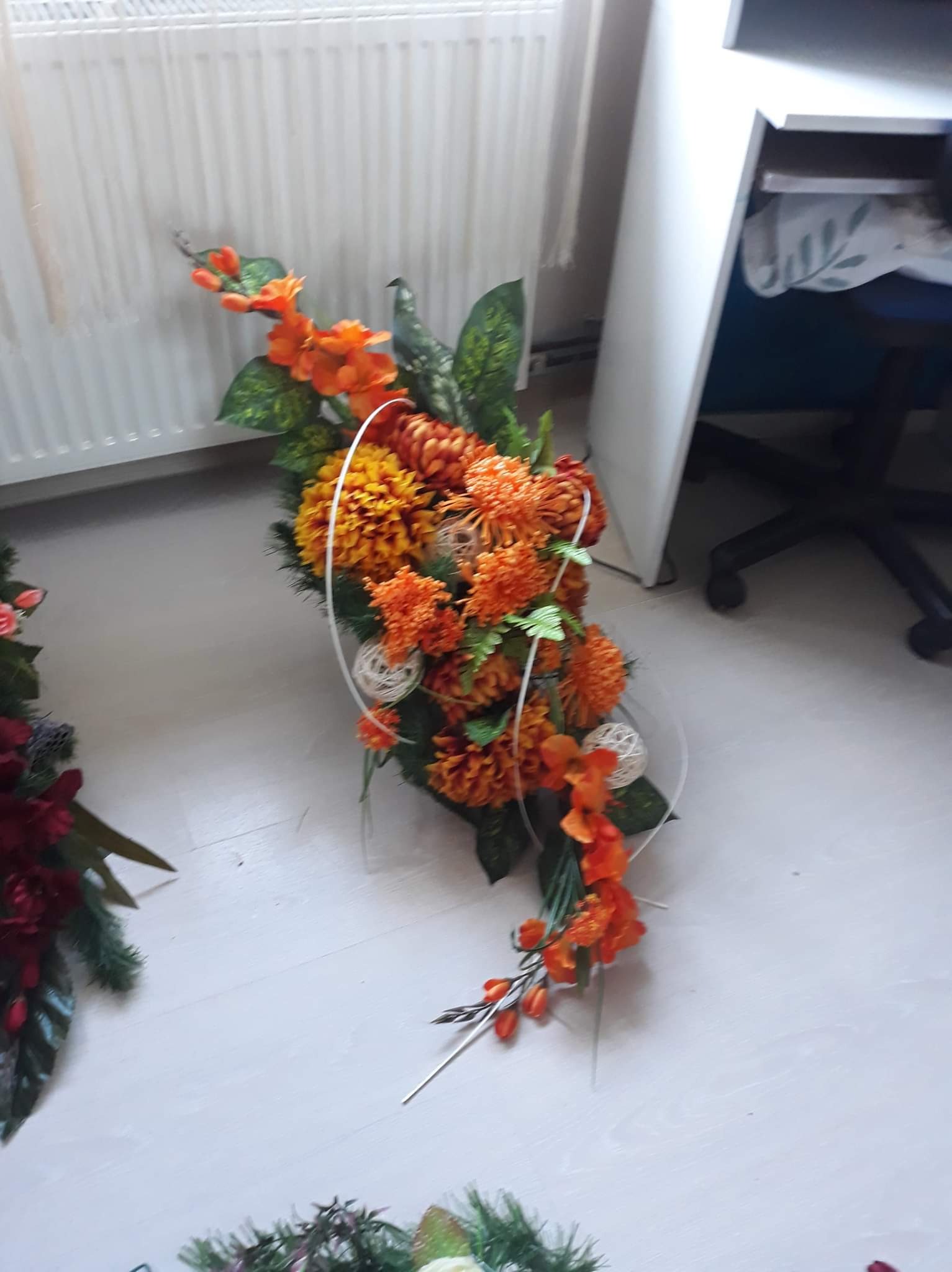 Pomarańczowo-żółty kosz prezentowy z kwiatów sztucznych i rattanowych kul, na jasnej podłodze obok grzejnika i biurka.