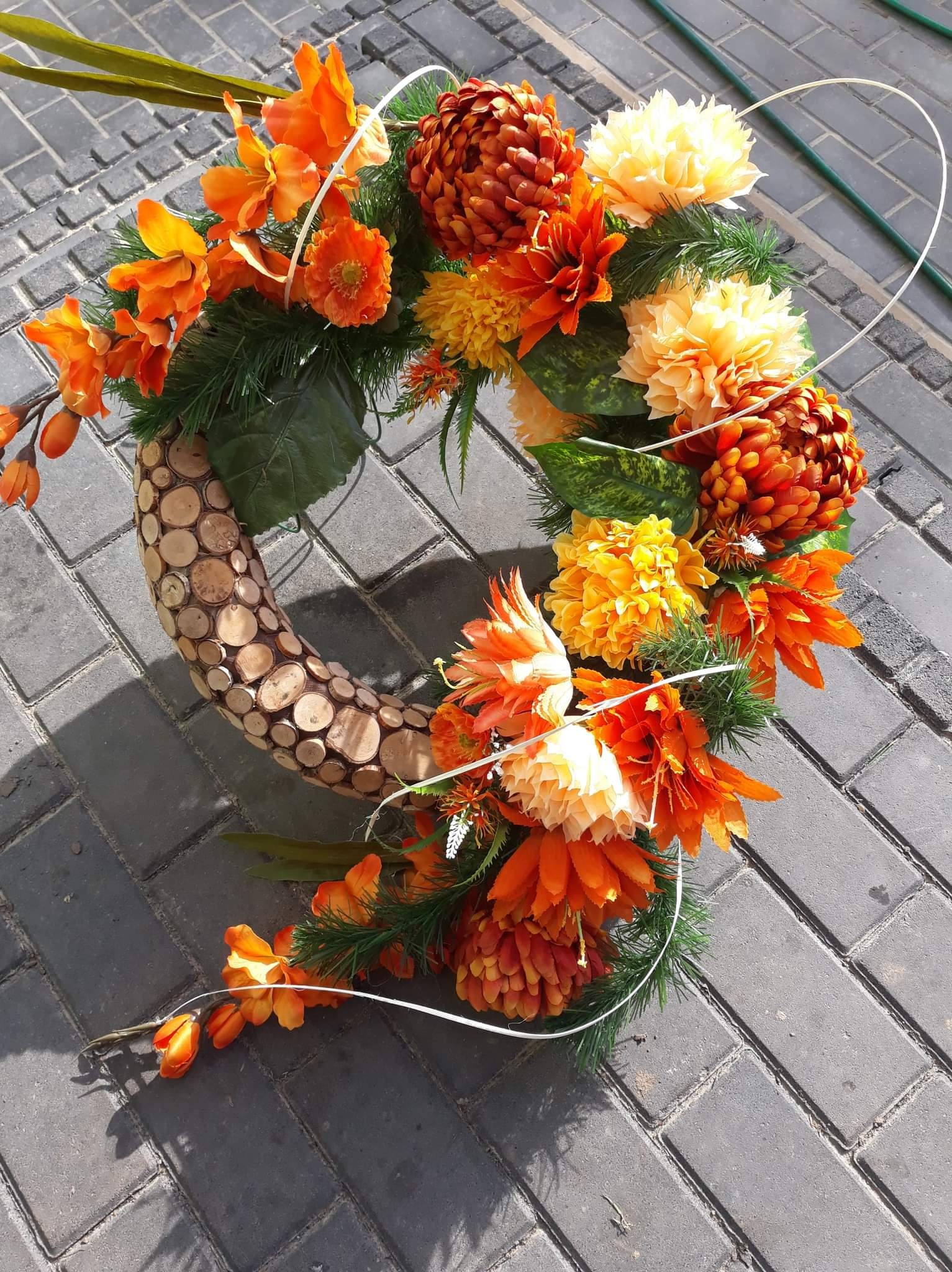 Dekoracyjny wieniec z pomarańczowych i żółtych kwiatów, z drewnianą podstawą z okrągłych plasterków drewna, leżący na szarej kostce brukowej.