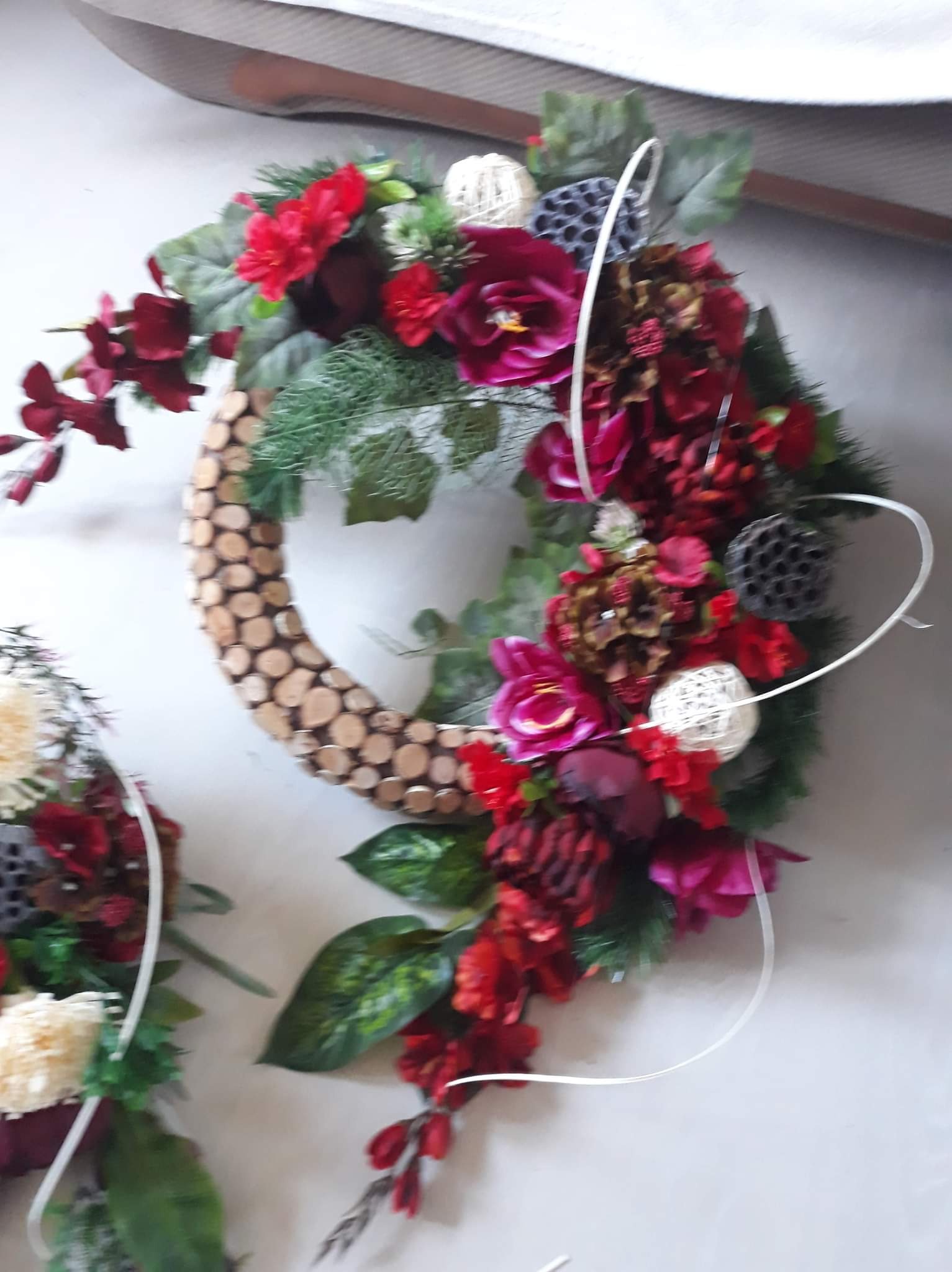 Dekoracyjny wieniec z czerwonych kwiatów, zielonych liści, drewnianych elementów i białych akcentów, leżący na jasnym tle, obok częściowo widocznego drugiego wieńca.