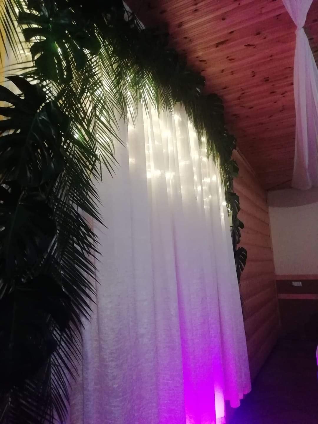 Dekoracja wnętrza z białymi zasłonami, oświetleniem LED i elementami roślinnymi, oświetlona fioletowym światłem, widoczny drewniany sufit i ściana.