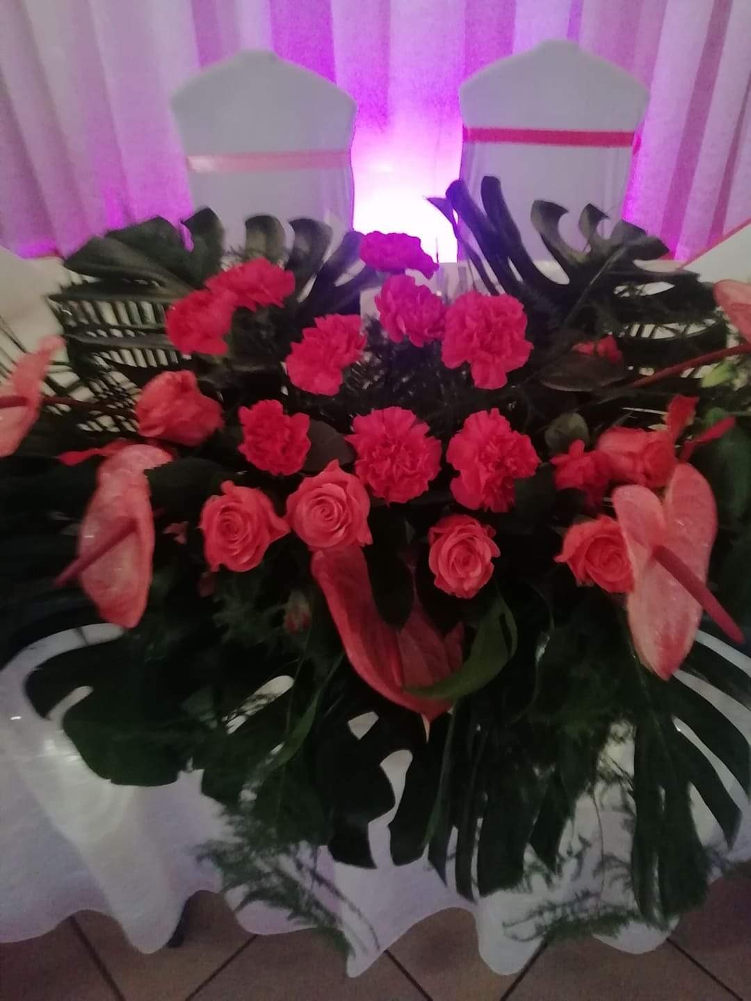 Kompozycja kwiatowa z czerwonych róż, anturium i liści monstery na białym obrusie, z krzesłami w tle oświetlonymi fioletowym światłem.