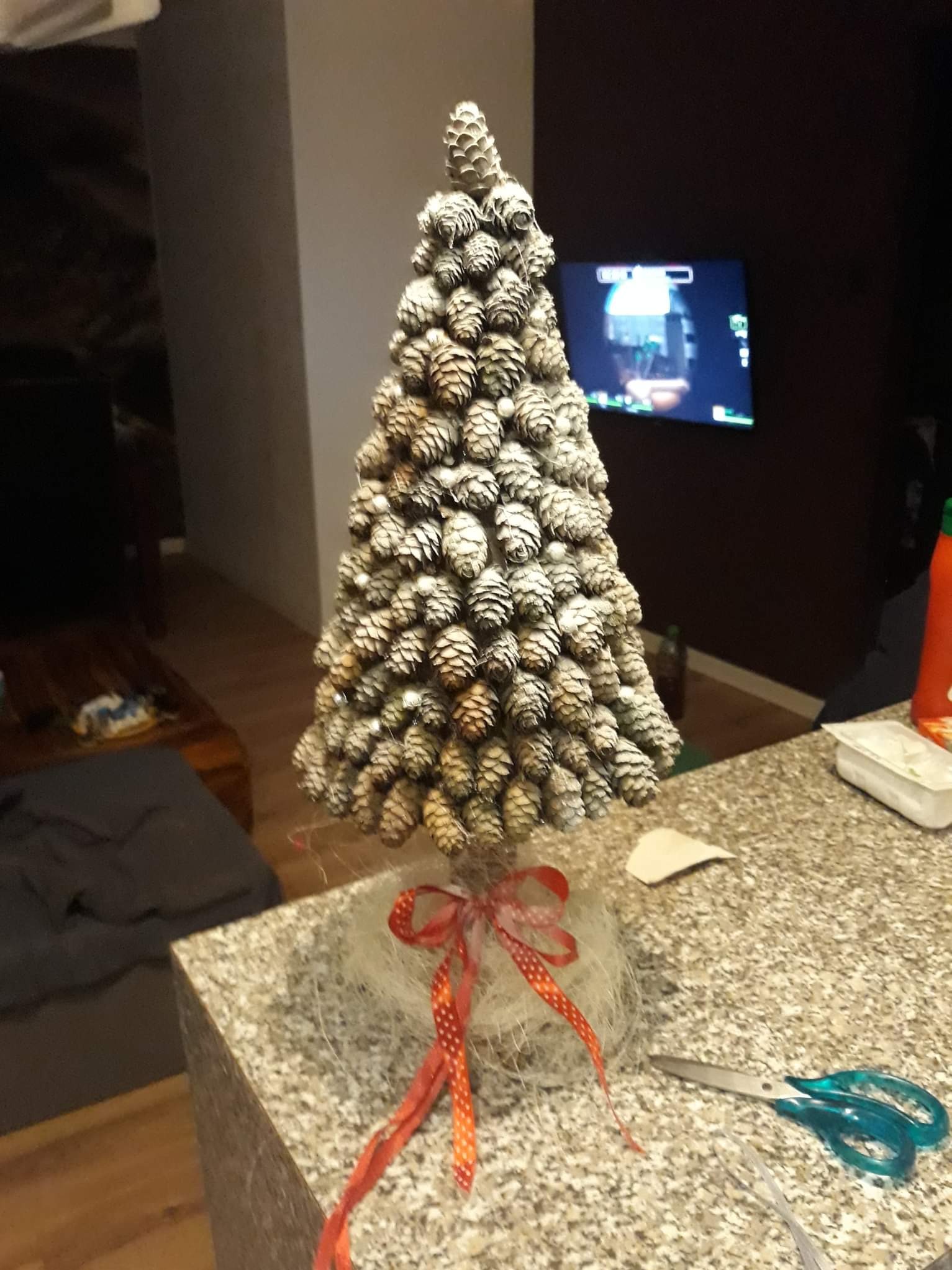 Dekoracyjna choinka z szyszek, ozdobiona czerwoną wstążką w kropki, stoi na blacie obok nożyczek z niebieskimi rączkami.