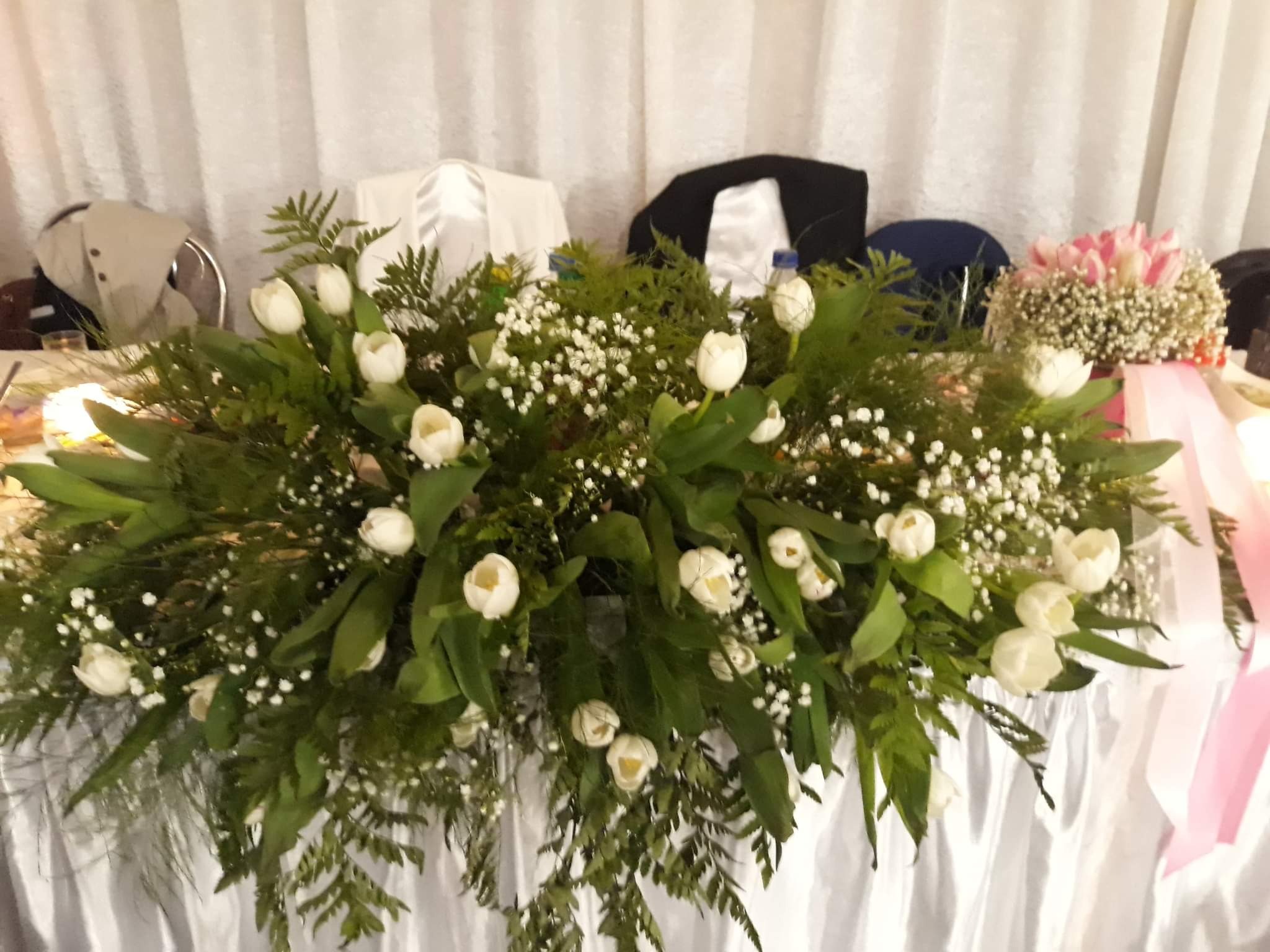 Dekoracja stołu z białych tulipanów, drobnych białych kwiatów i zielonych liści na tle białej zasłony i krzeseł.